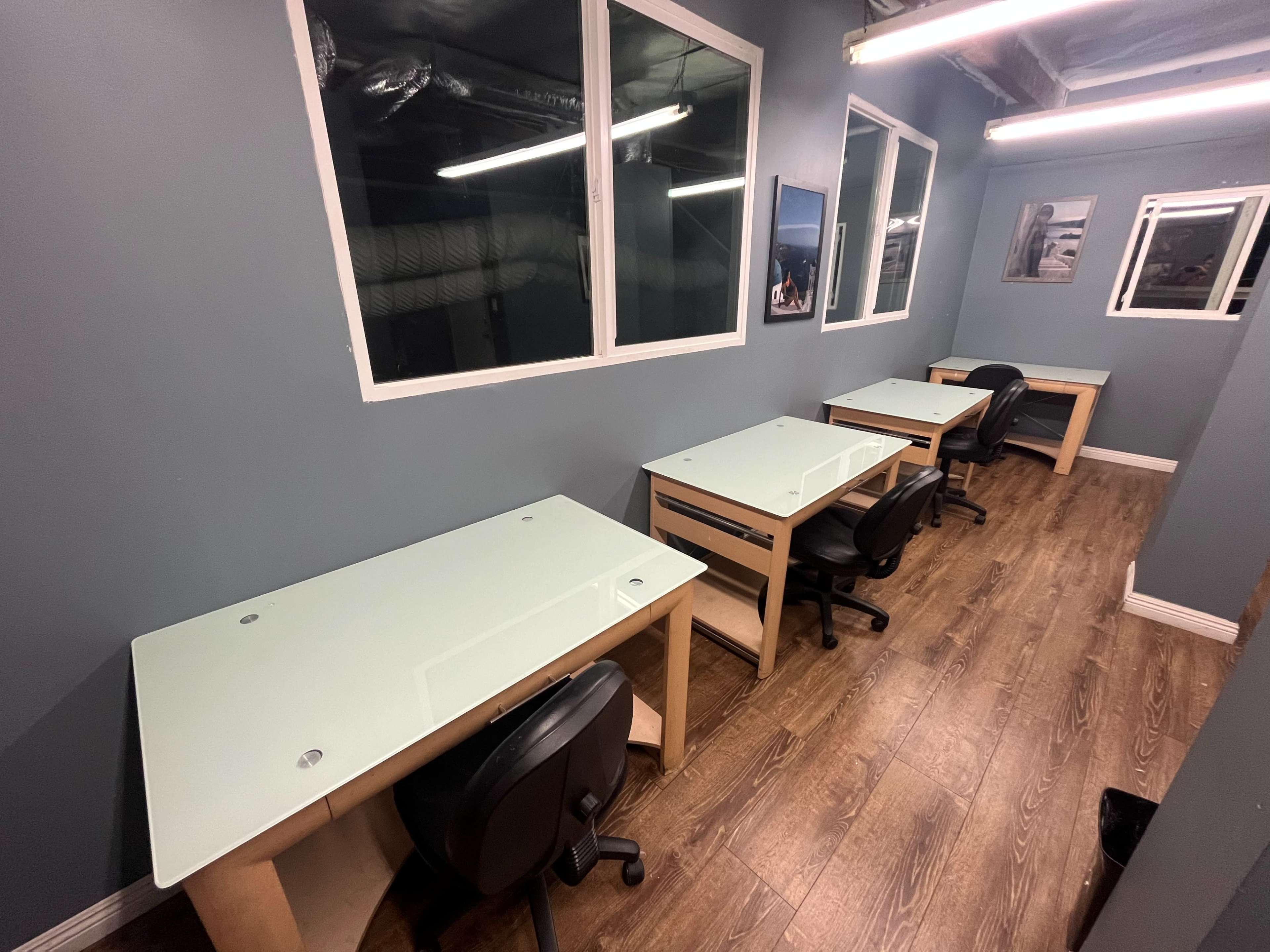 The image shows a row of four glass-top desks with black office chairs in a room with gray walls and several windows.