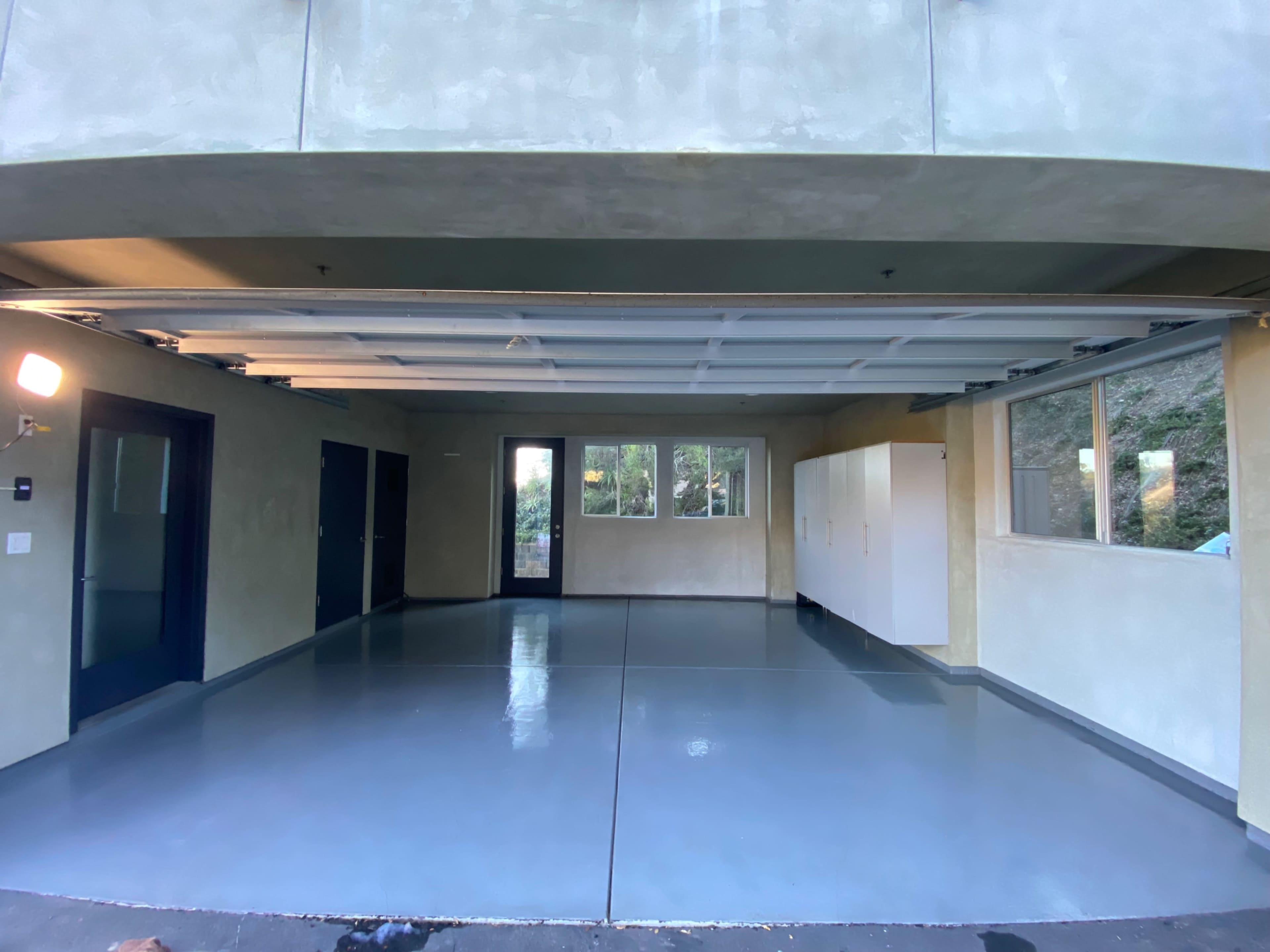 The image shows a spacious, empty garage with a grey floor and walls, featuring large windows and white storage cabinets.