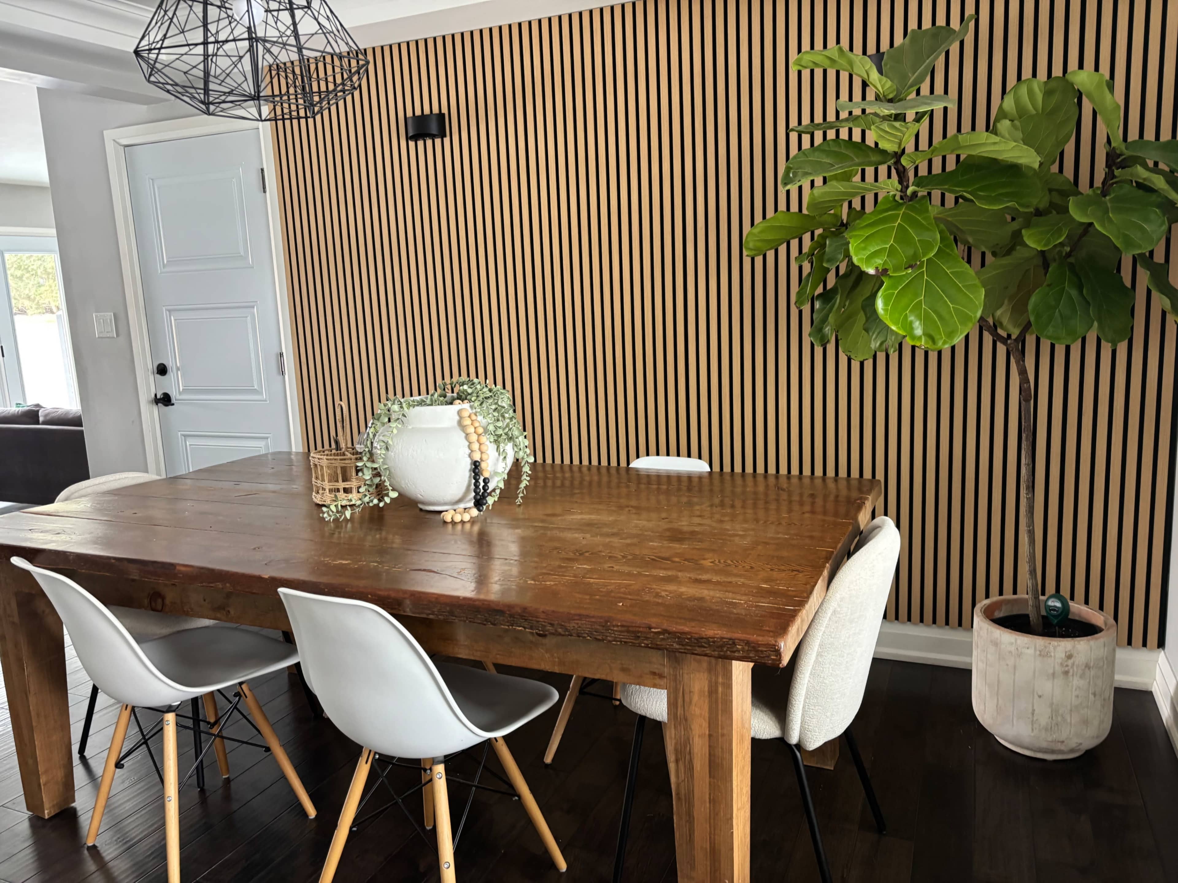 A wooden dining table with four chairs is placed in a room featuring a striped wooden accent wall and a potted fiddle leaf fig plant.