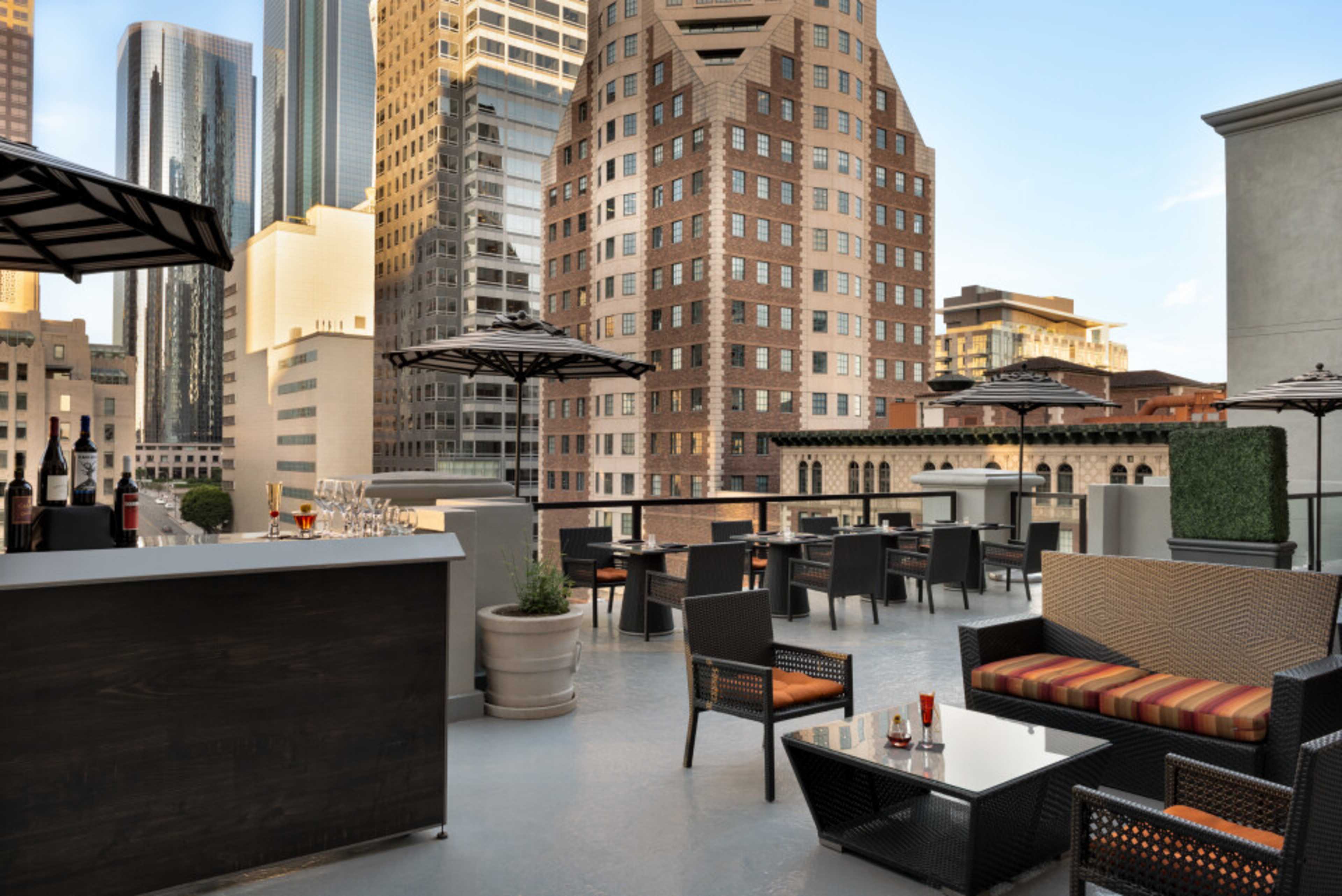 The image shows a rooftop terrace with seating, an outdoor bar area, and tall buildings in the background.