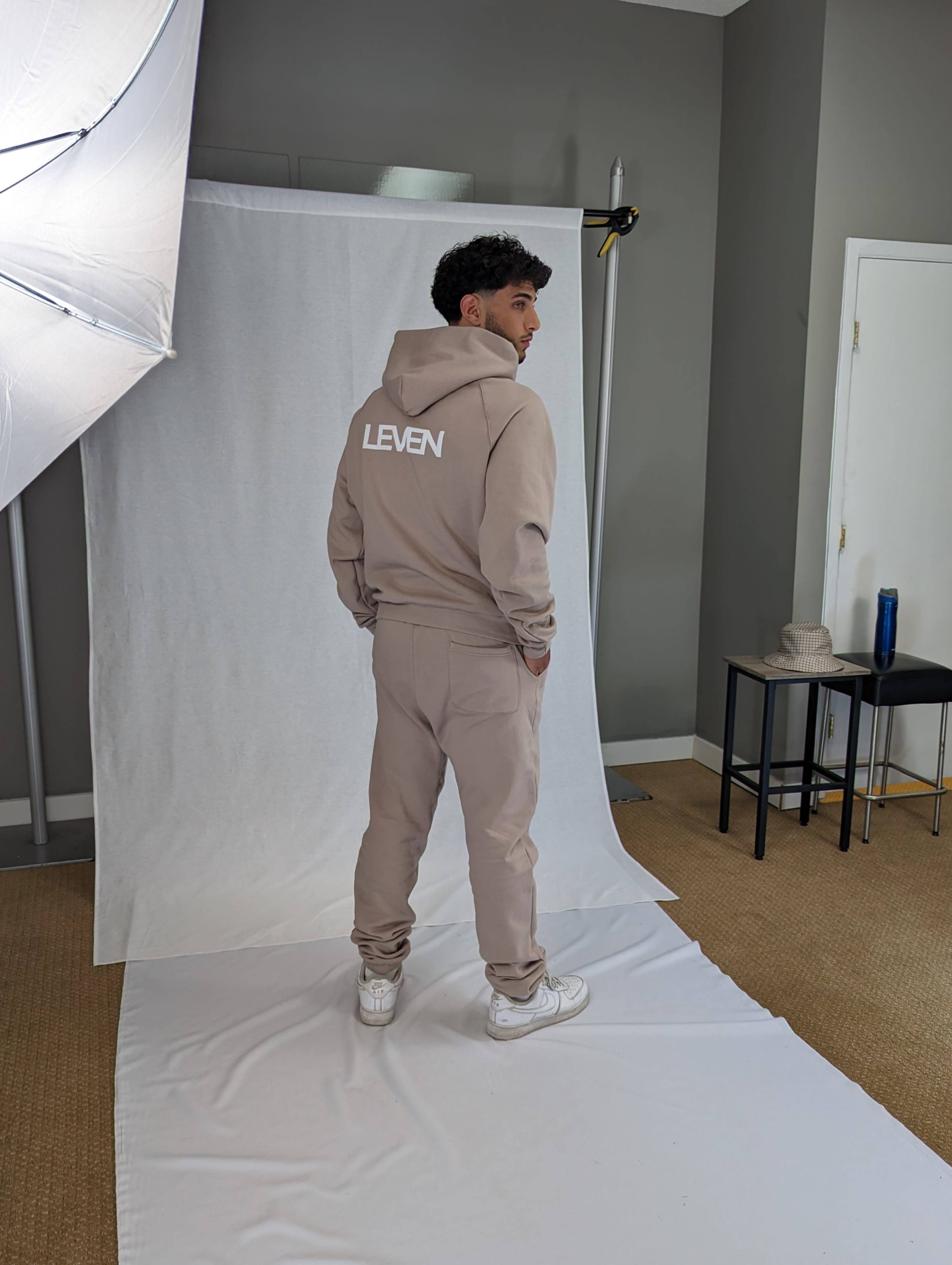 A person stands facing away from the camera in a studio, wearing a beige hoodie and matching pants with the word "LEVEN" printed on the back.