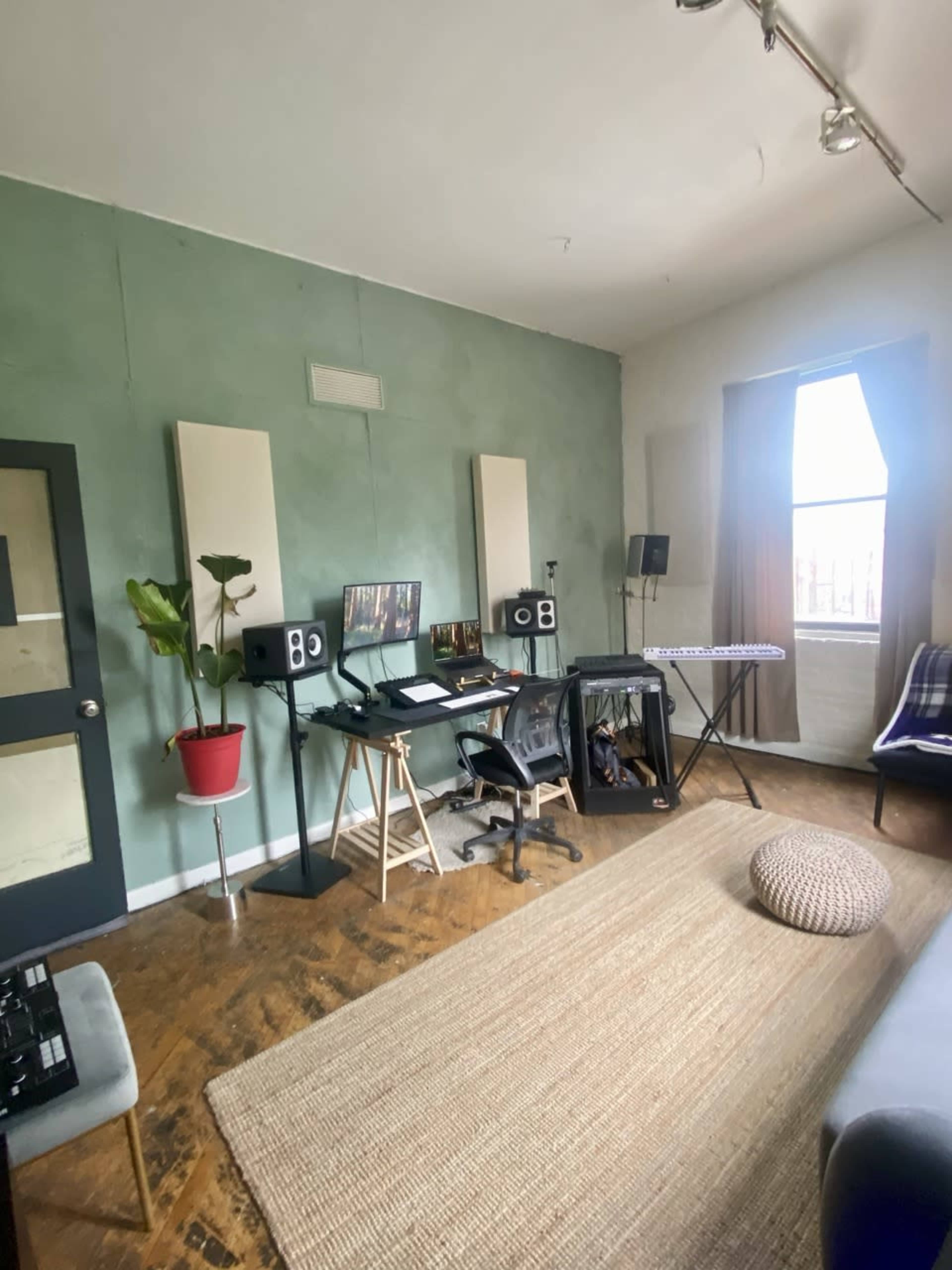The image shows a music studio with a desk, computer, monitors, keyboard, and acoustic panels on the walls, featuring a plant in a red pot and a large rug on the floor.