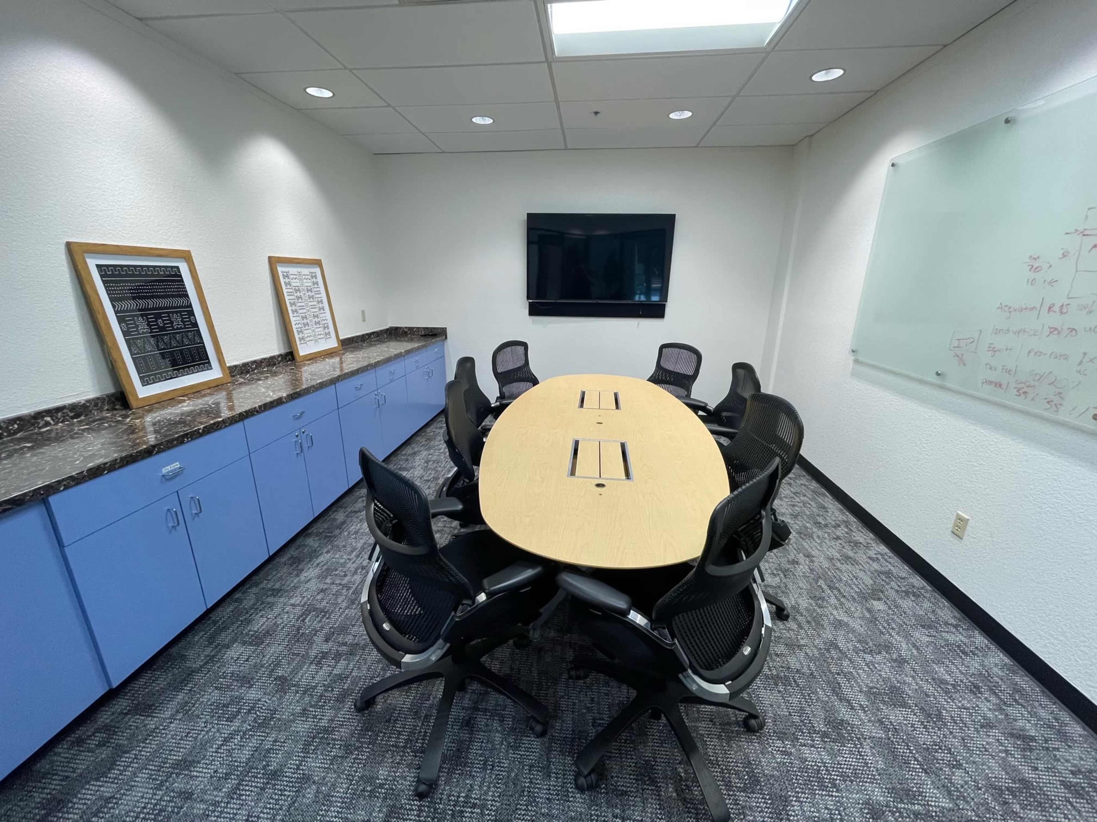 A conference room features a large oval table surrounded by black office chairs, with a television and whiteboards on the walls.