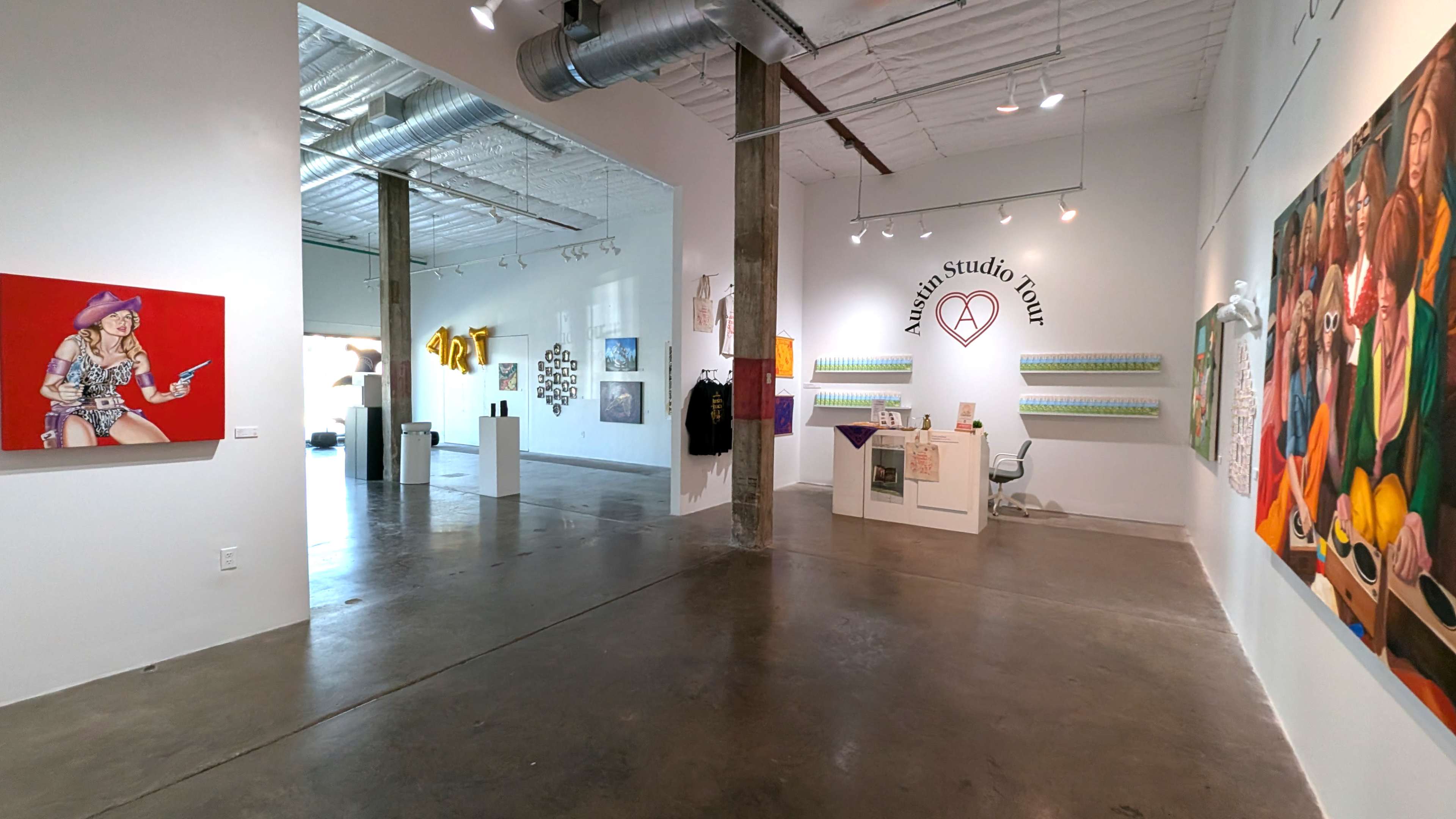 The image shows an art gallery with a spacious interior featuring multiple artworks displayed on the walls and a reception area with a desk.