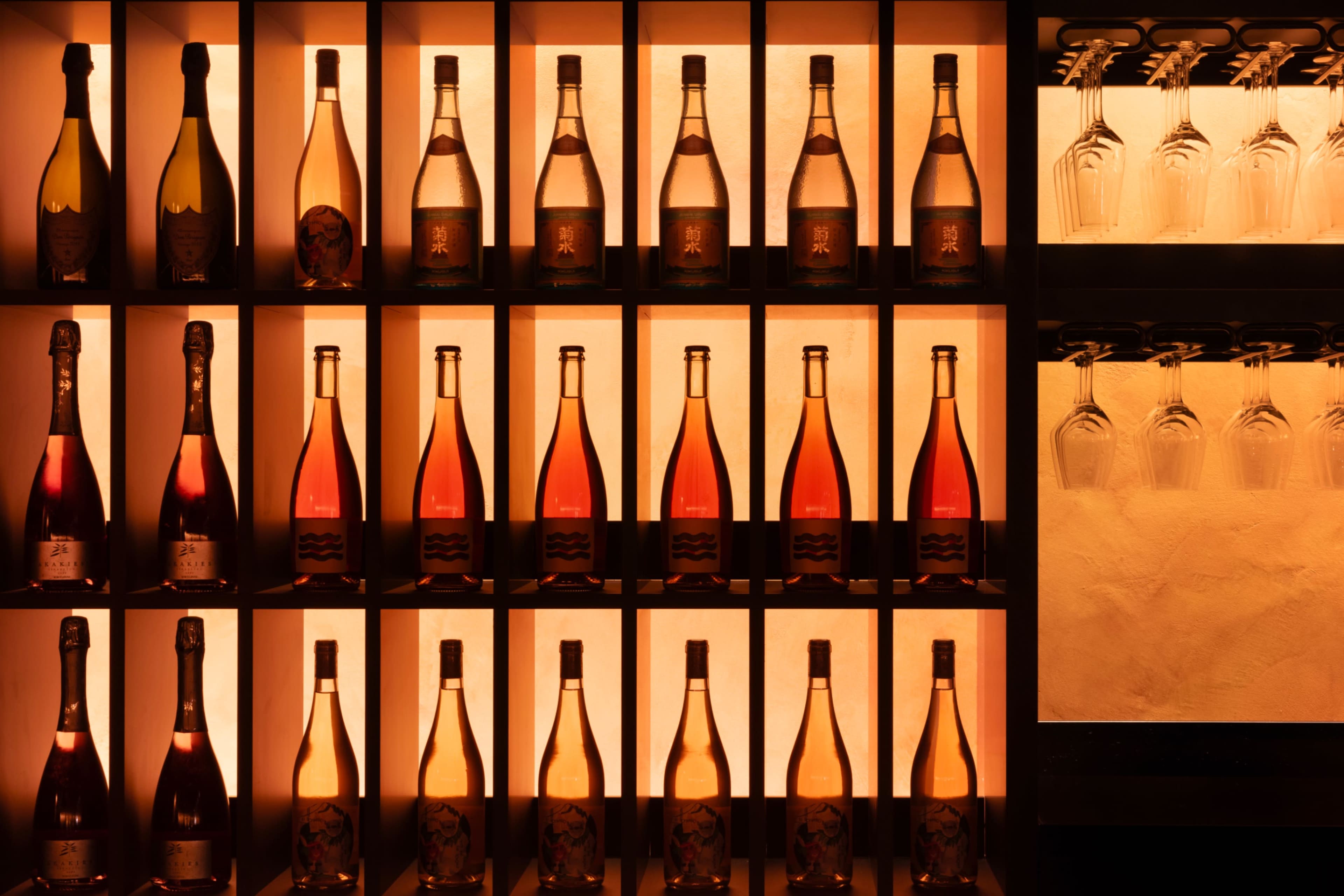 A wall display showcases various bottles of wine and champagne, illuminated by soft orange lighting, with wine glasses hanging above.