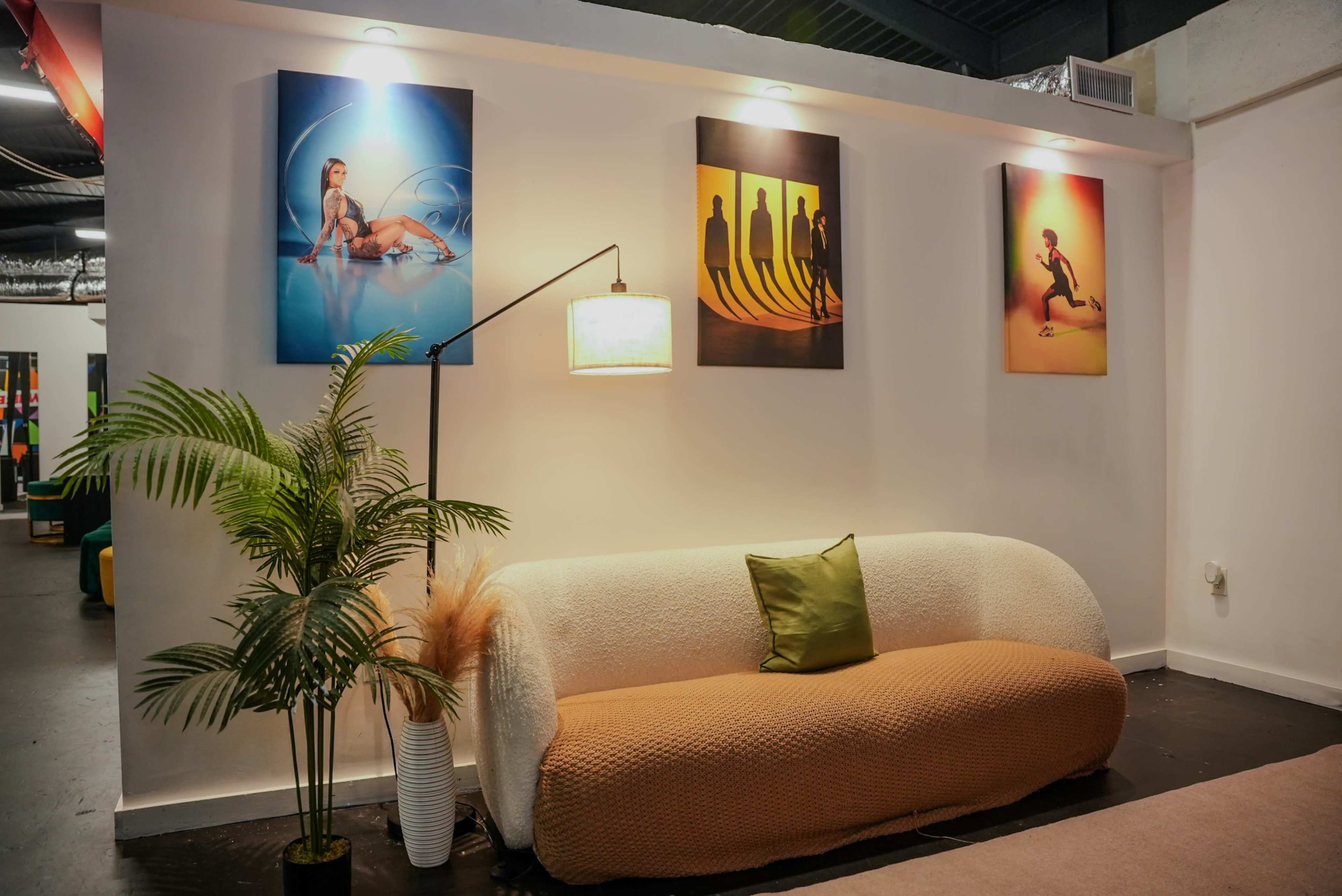 The image shows a modern interior space featuring a white sofa with an orange accent, a floor lamp, a potted plant, and three colorful framed artworks on the wall.