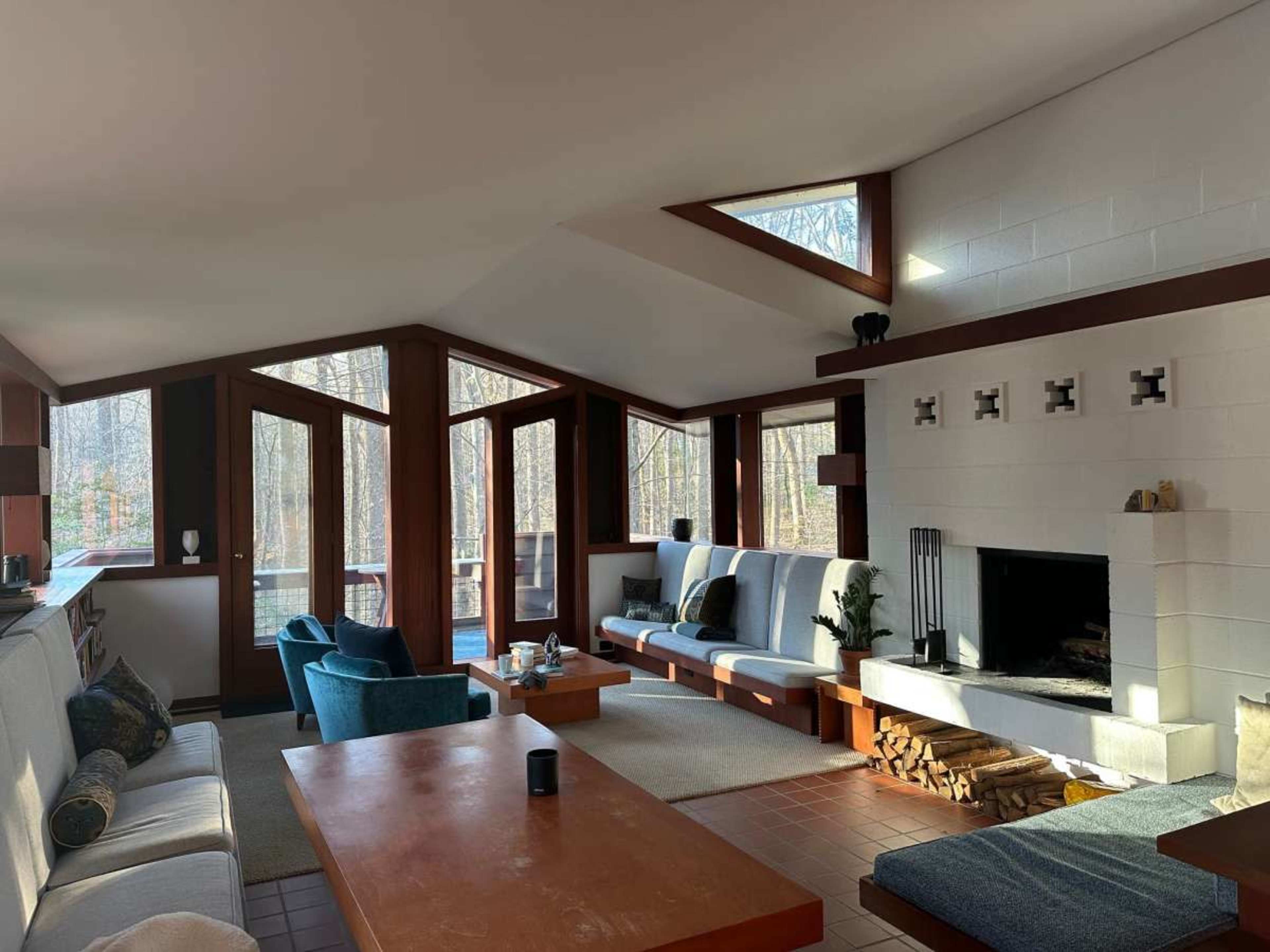 A modern living room with large windows overlooking a wooded area, featuring a fireplace, comfortable seating, and wooden furniture.
