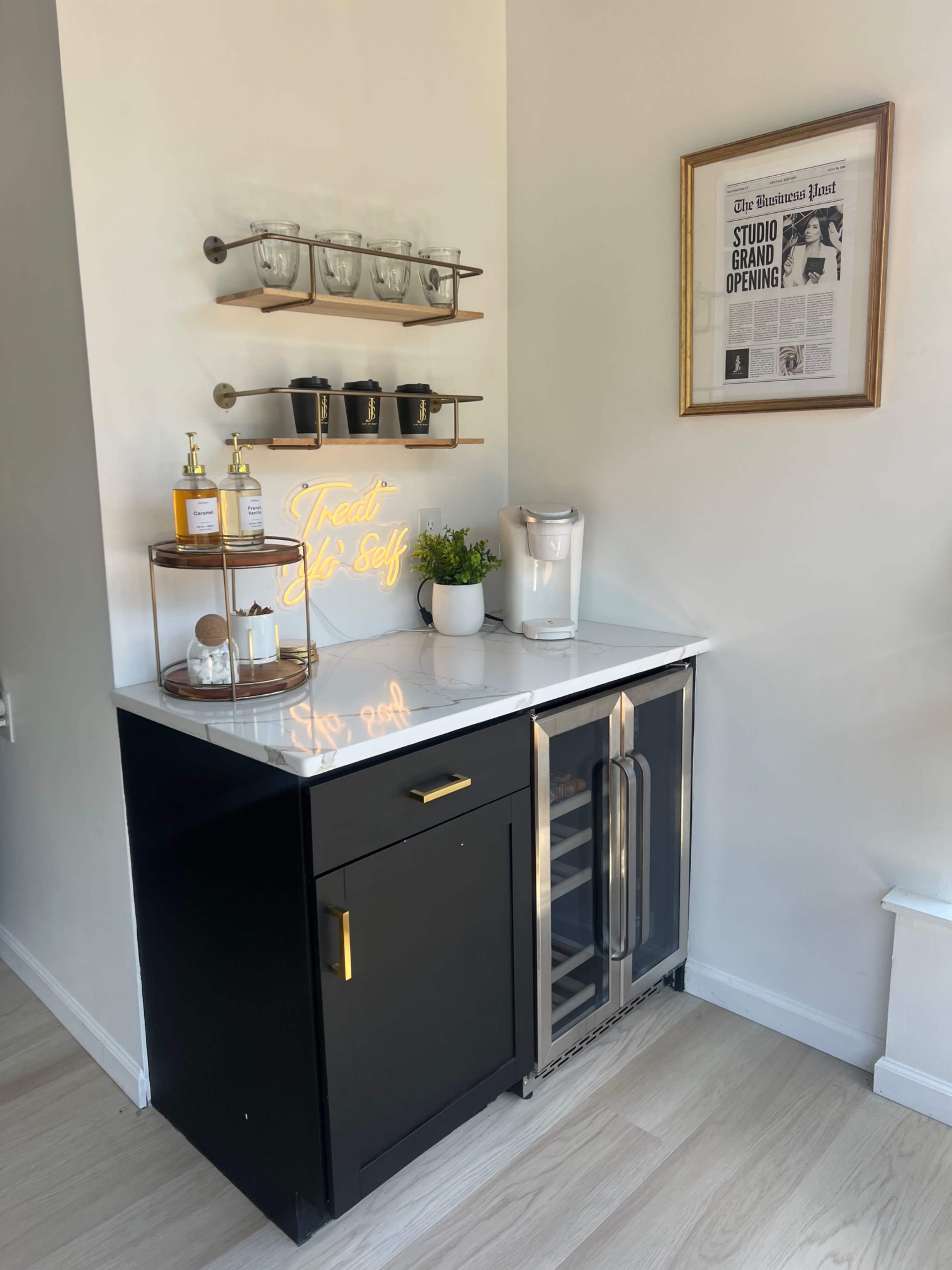 A modern kitchenette featuring a black cabinet with a marble countertop, a mini-fridge, shelving with glassware, and a sign that says "Treat Yo' Self."