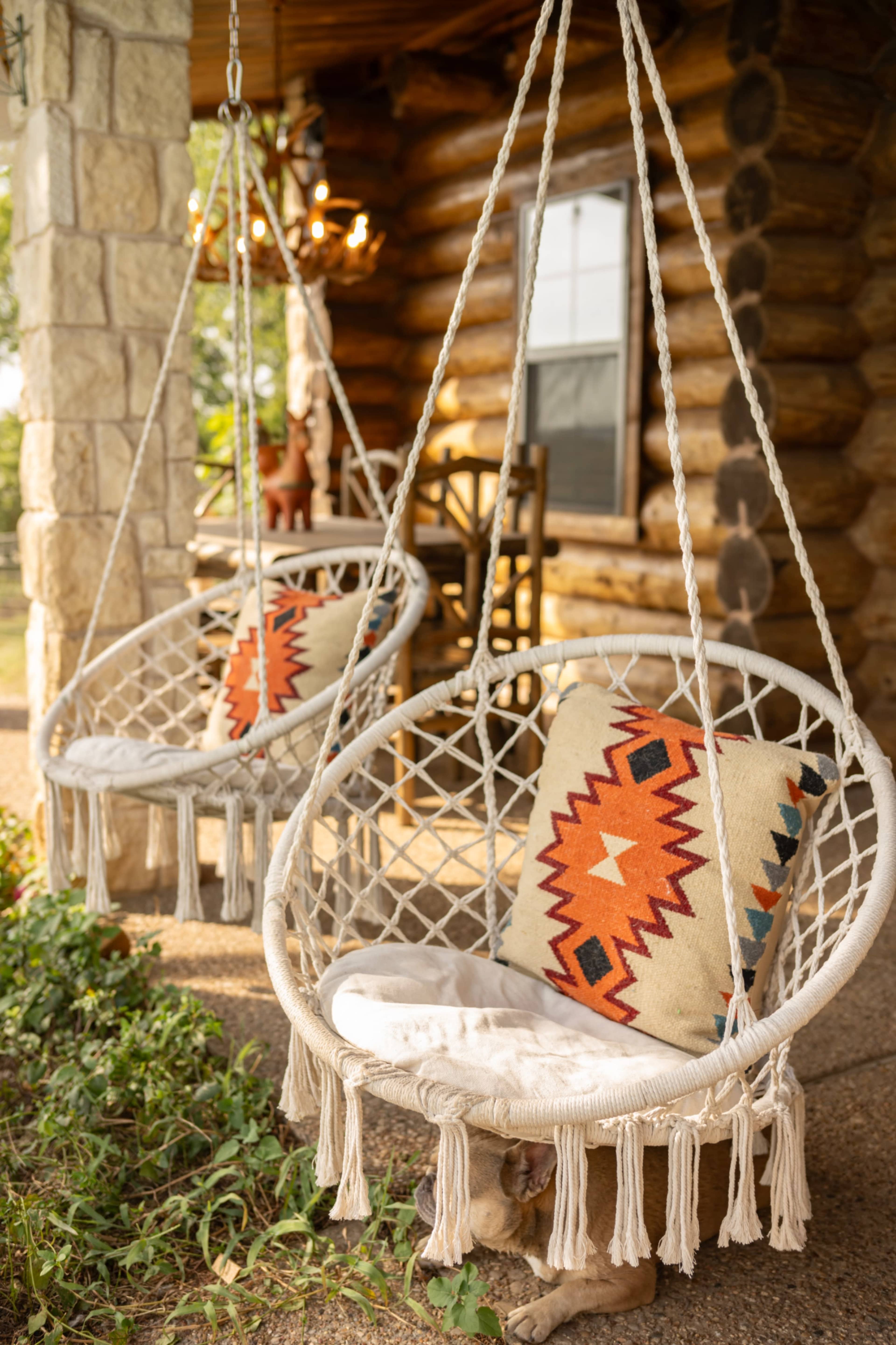 Two hanging macrame chairs with decorative cushions are positioned on a porch beside a log cabin.