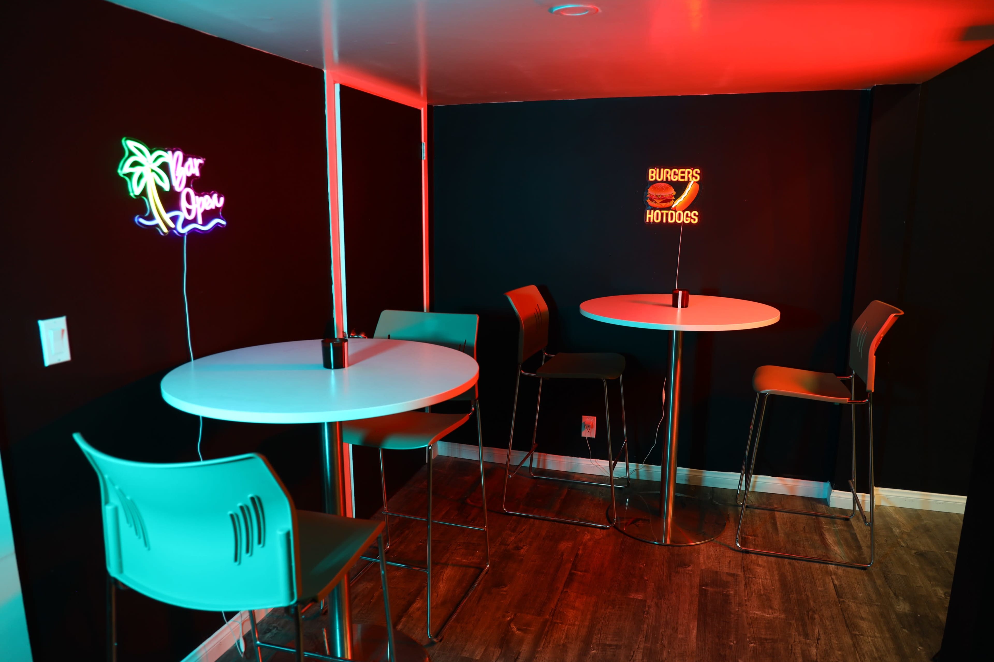 The image shows a small indoor space with two high tables and chairs, illuminated by neon signs that read "Your Space" and "BURGERS HOTDOGS."