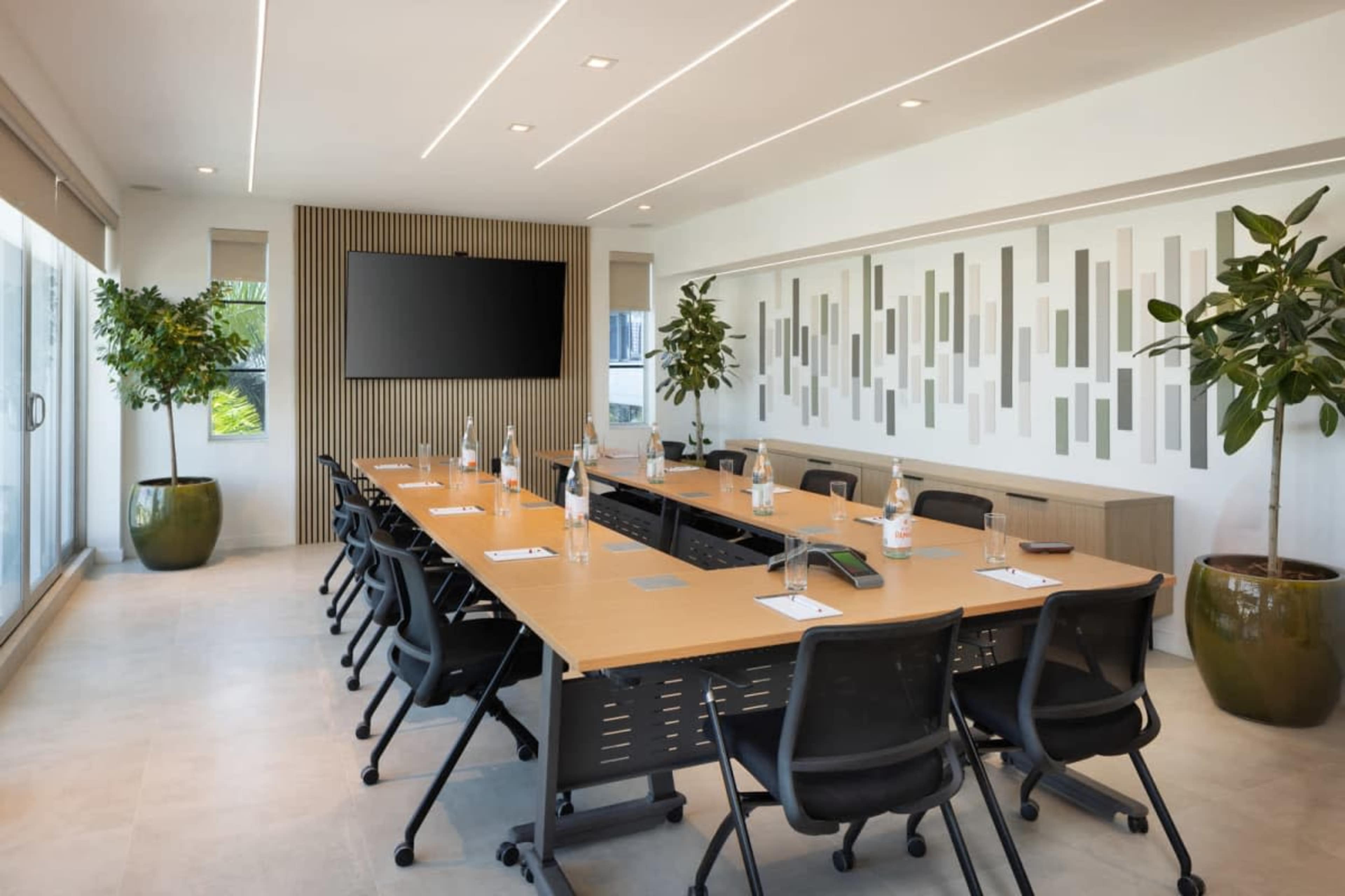 A modern conference room features a long wooden table surrounded by black chairs, with large plants in decorative pots and a wall-mounted screen.