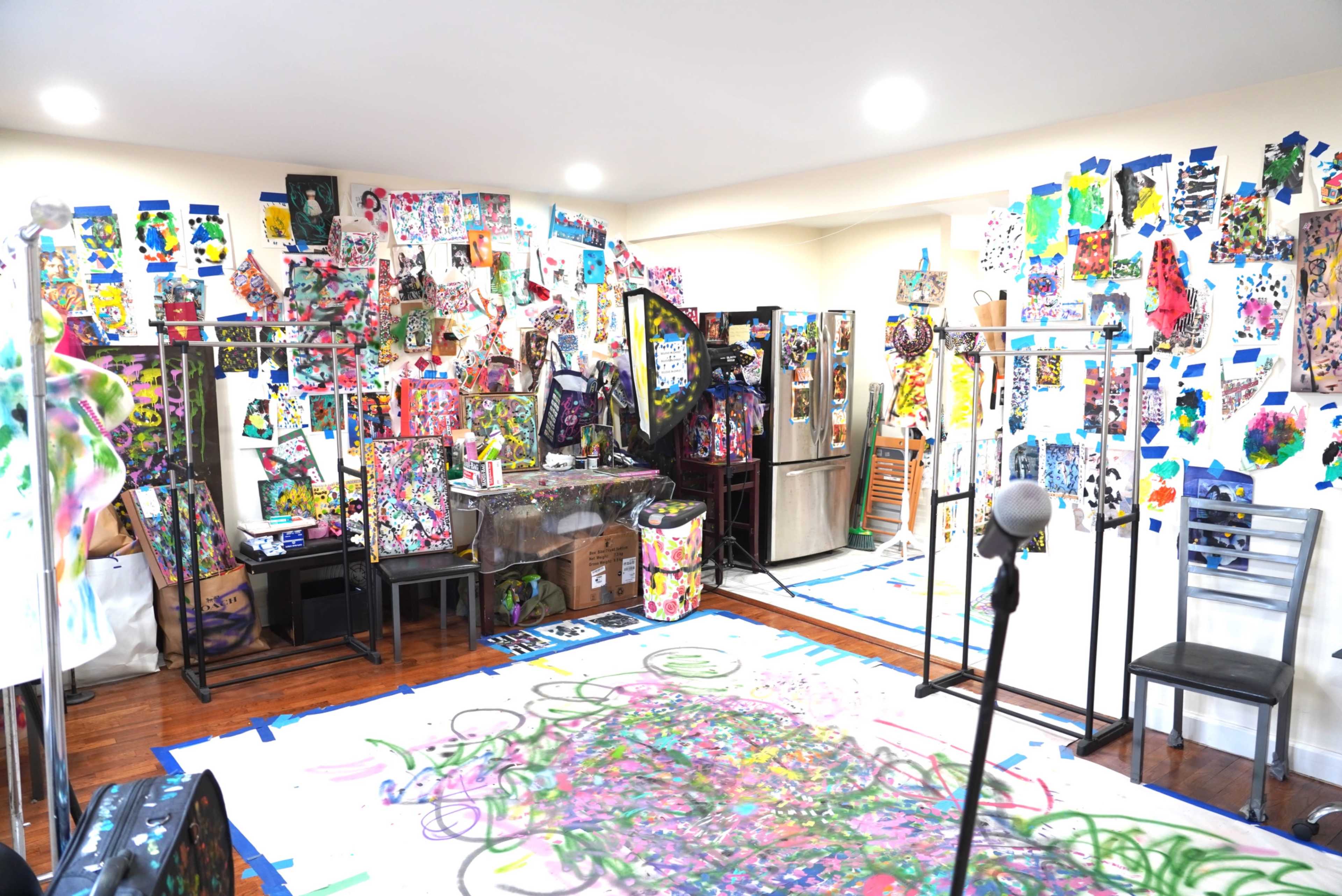 The image shows a vibrant art studio filled with colorful artworks and decorations covering the walls, along with various art supplies and furniture arranged throughout the space.