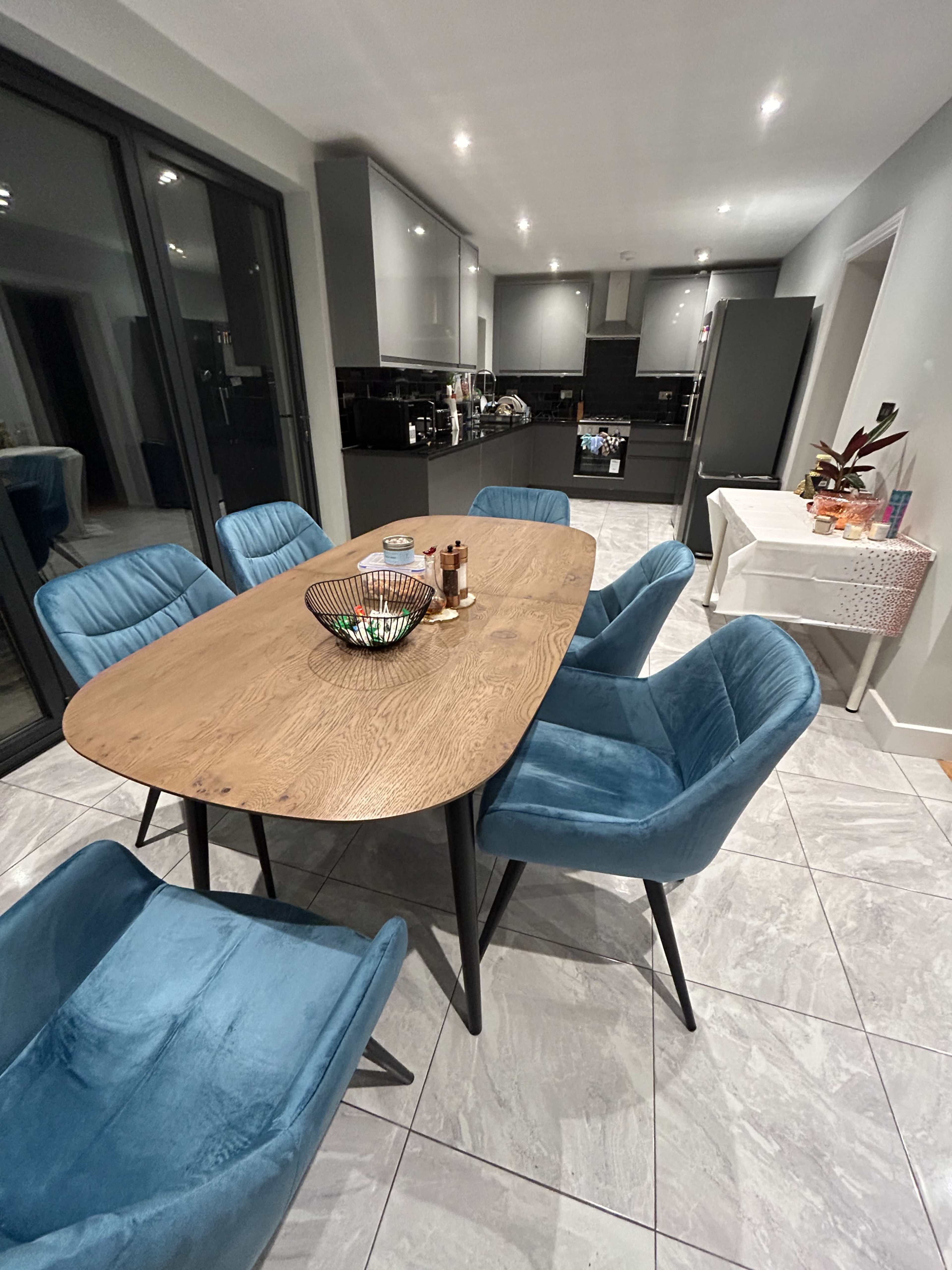 A modern dining area with a large wooden table surrounded by blue upholstered chairs and a kitchen visible in the background.