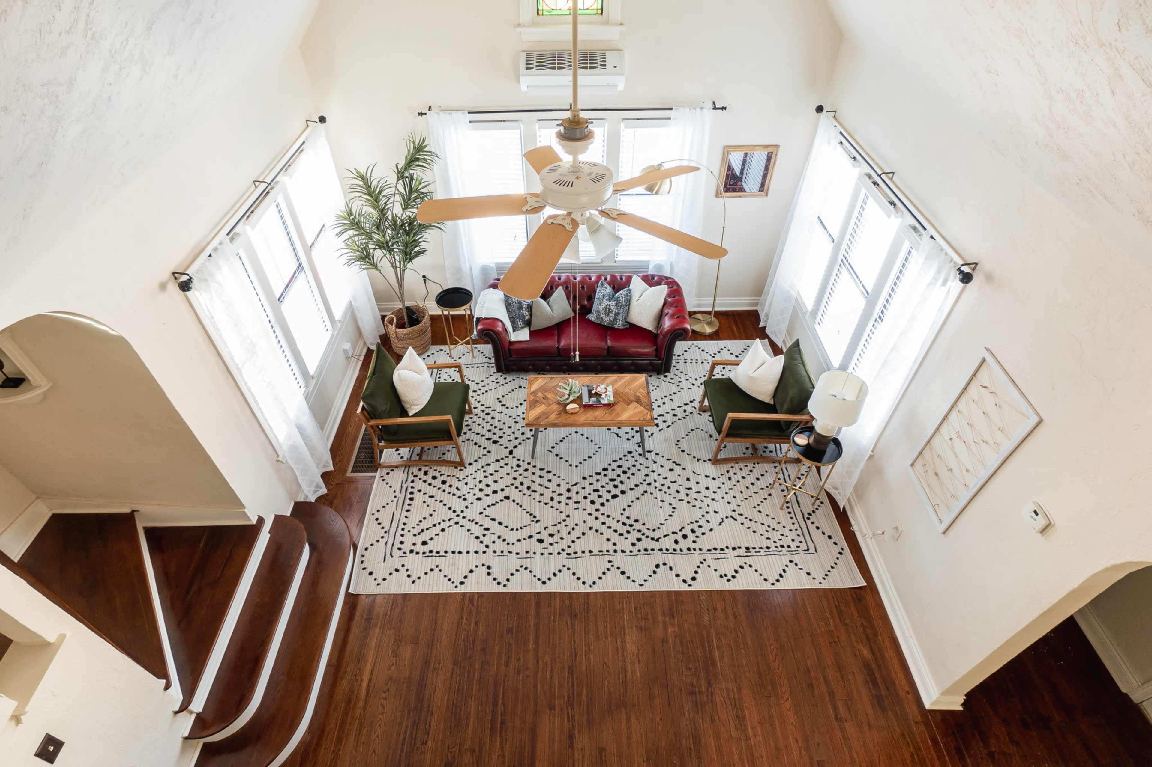 A bright living room with a red sofa, green chairs, a wooden coffee table, and a patterned area rug, all arranged under a ceiling fan.