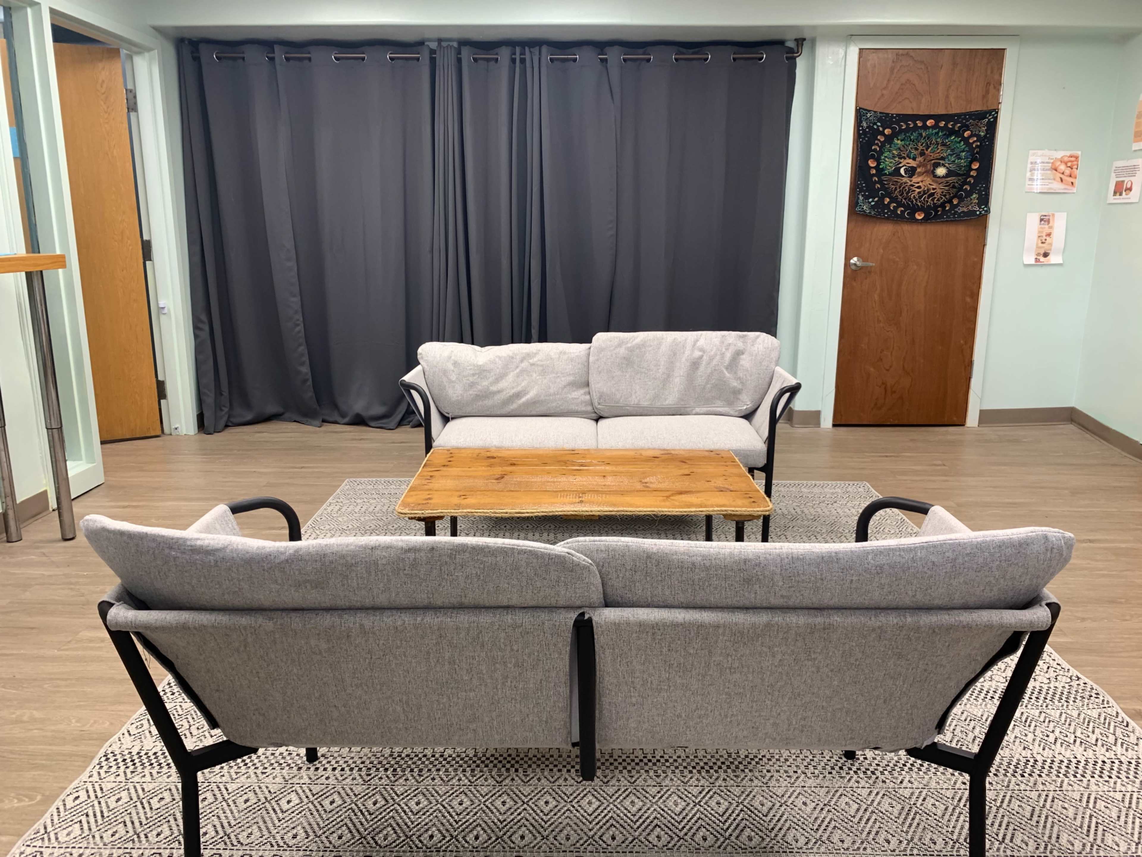 The image shows a seating area with two gray sofas facing a wooden coffee table, placed on a patterned rug, in a room with curtain-draped windows and doors.