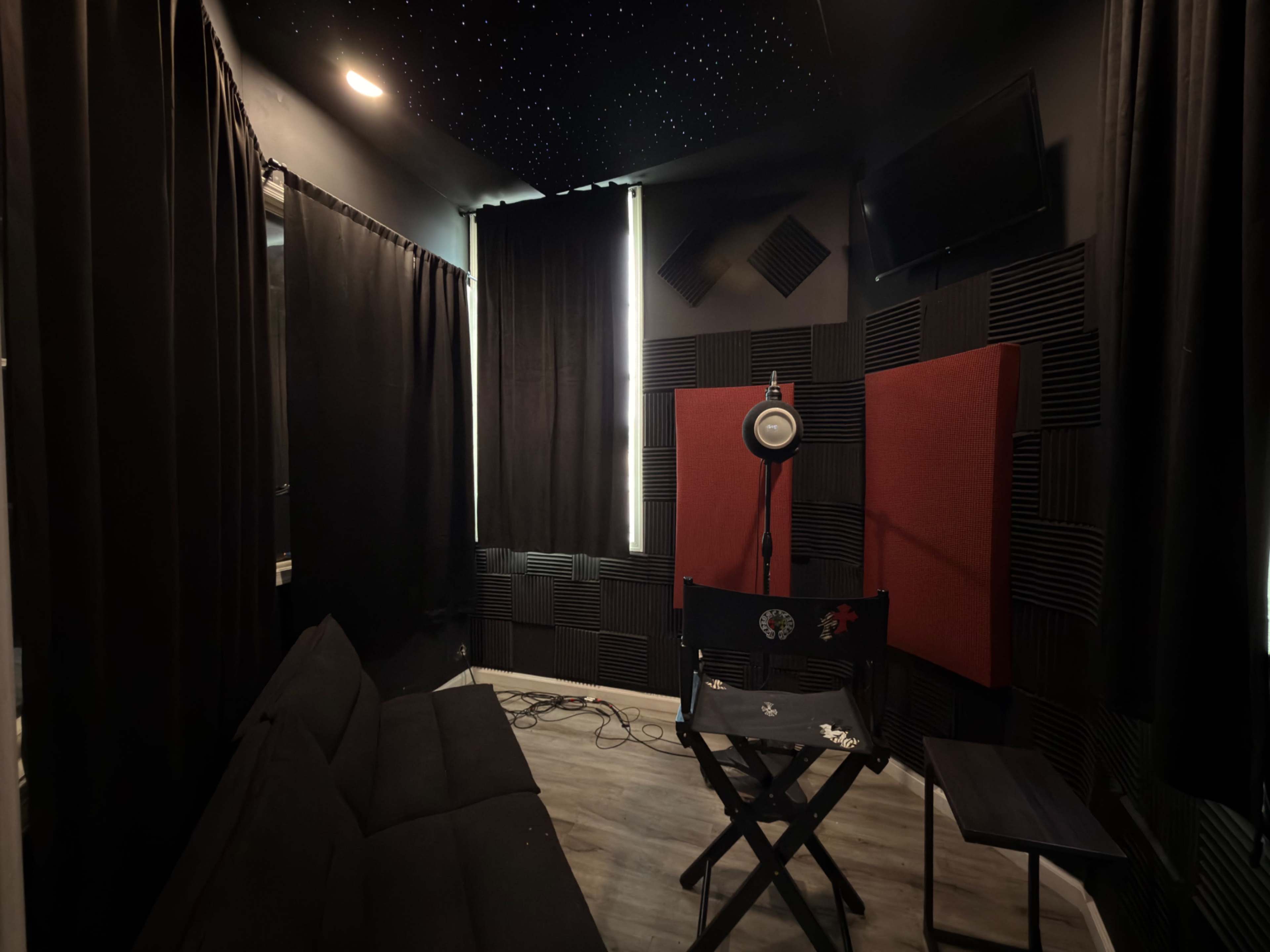 The image shows a small recording studio with soundproofing panels, a director's chair, a microphone setup, and black curtains covering the windows.