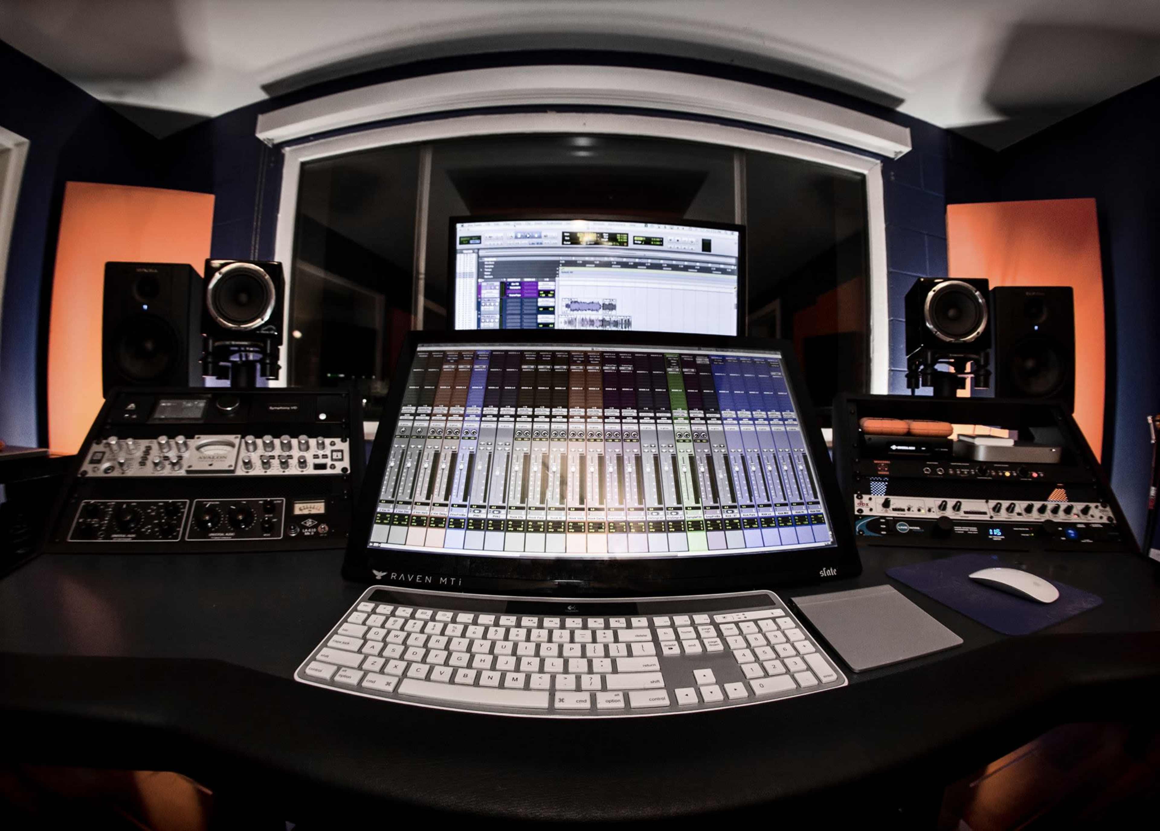 A modern audio recording studio with a large mixing console, computer monitors, and studio speakers arranged around a central workstation.