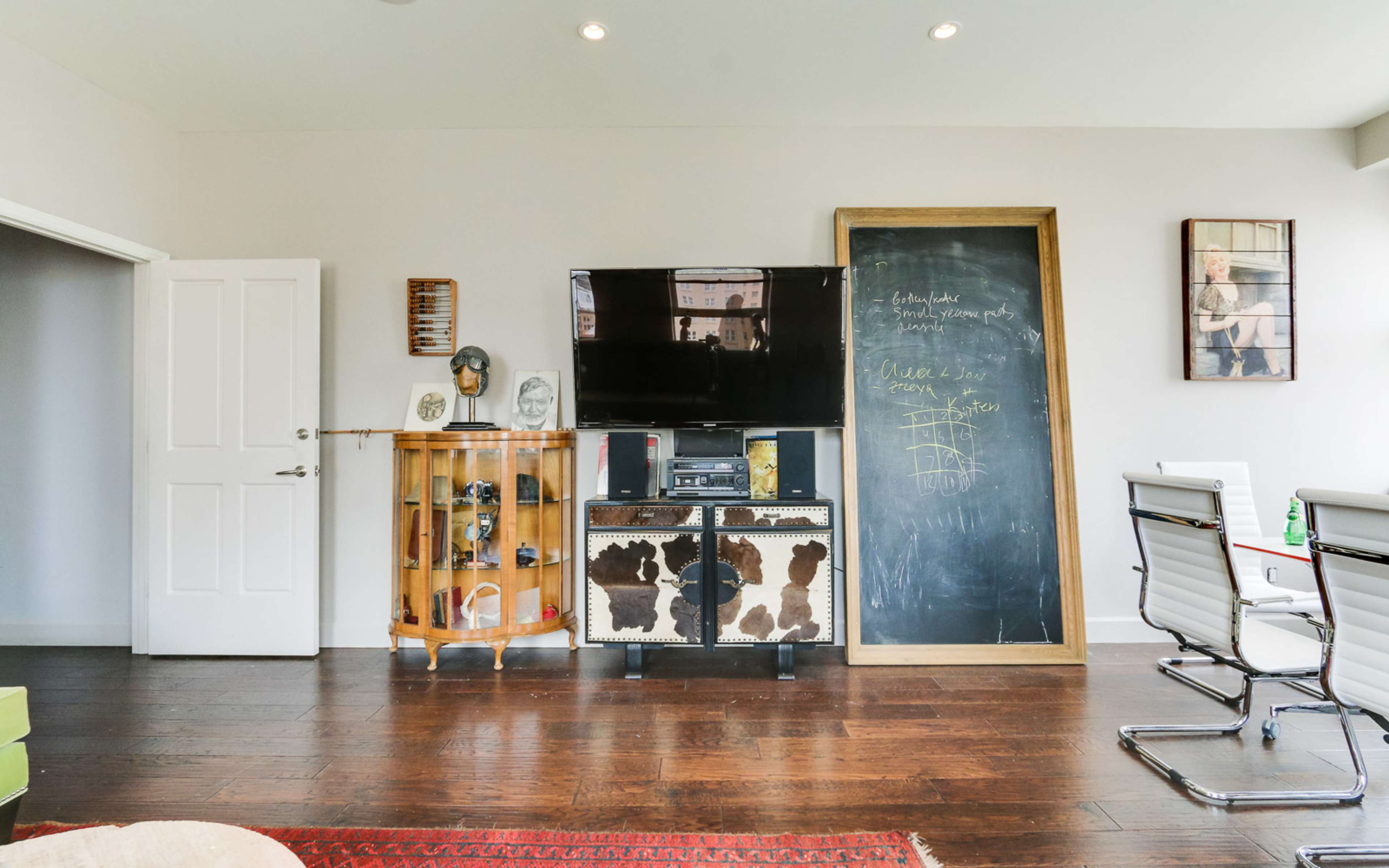 A modern office space featuring a large flat-screen TV, a decorative cabinet, a chalkboard, and a glass table with metal chairs.