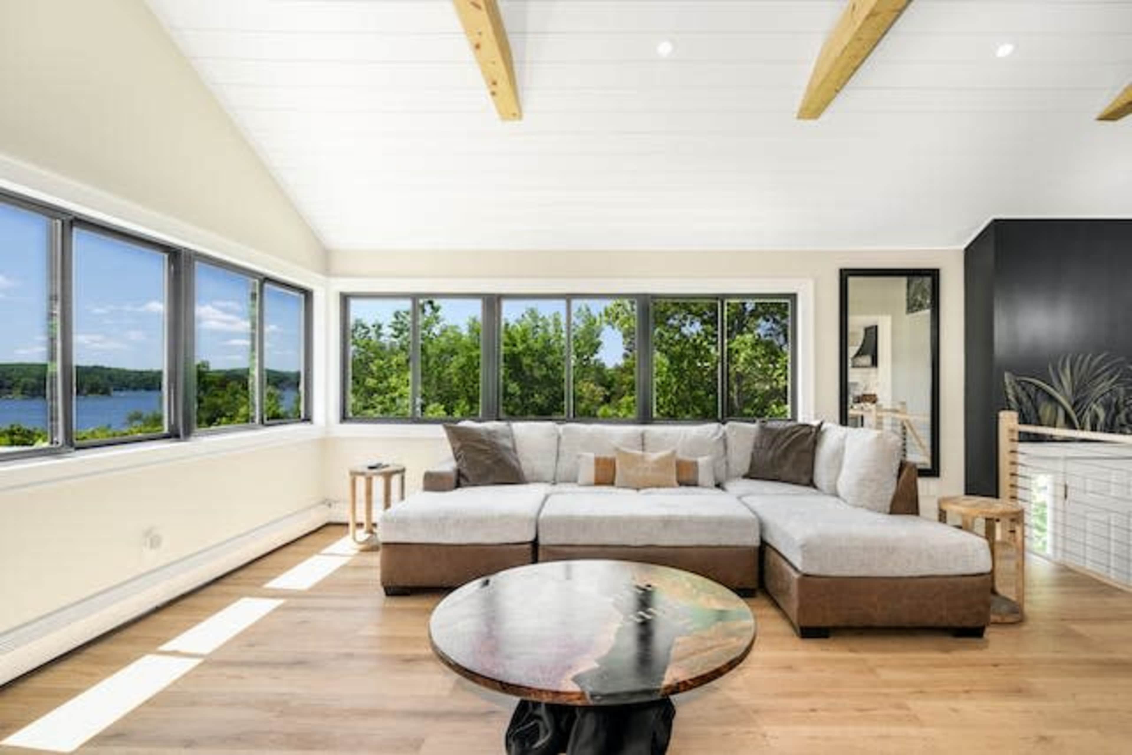 A spacious living room features a large sectional sofa, panoramic windows with lake views, and wooden beams on the ceiling.