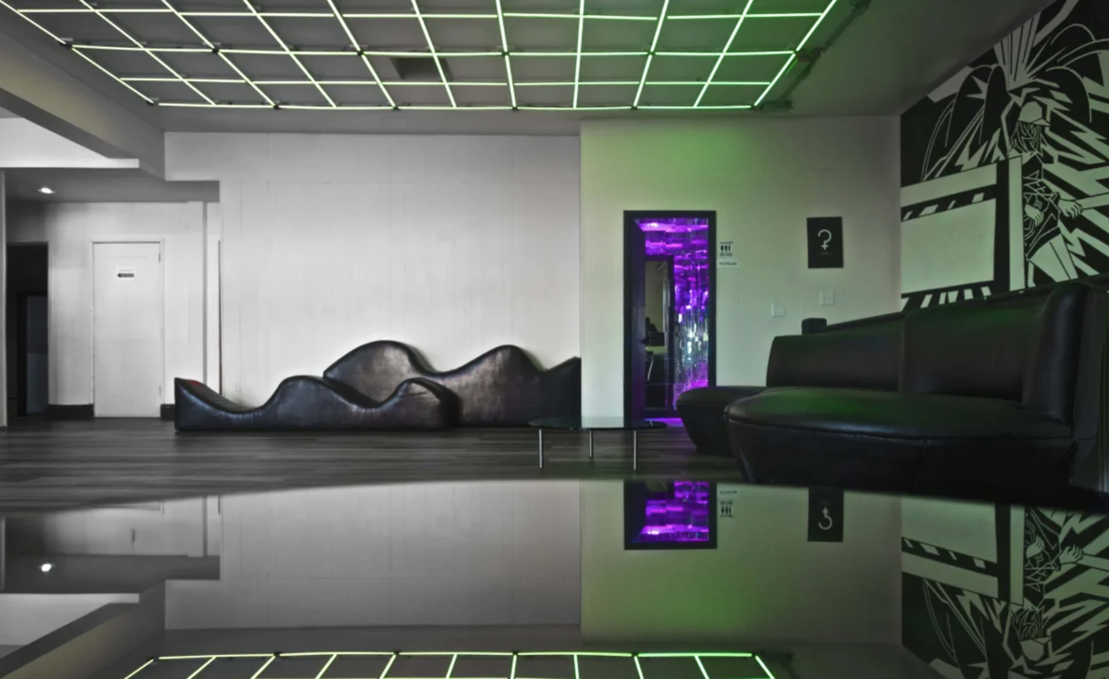 A modern interior with a reflective floor, featuring a curved black sofa and a glowing purple mirror at the far end.