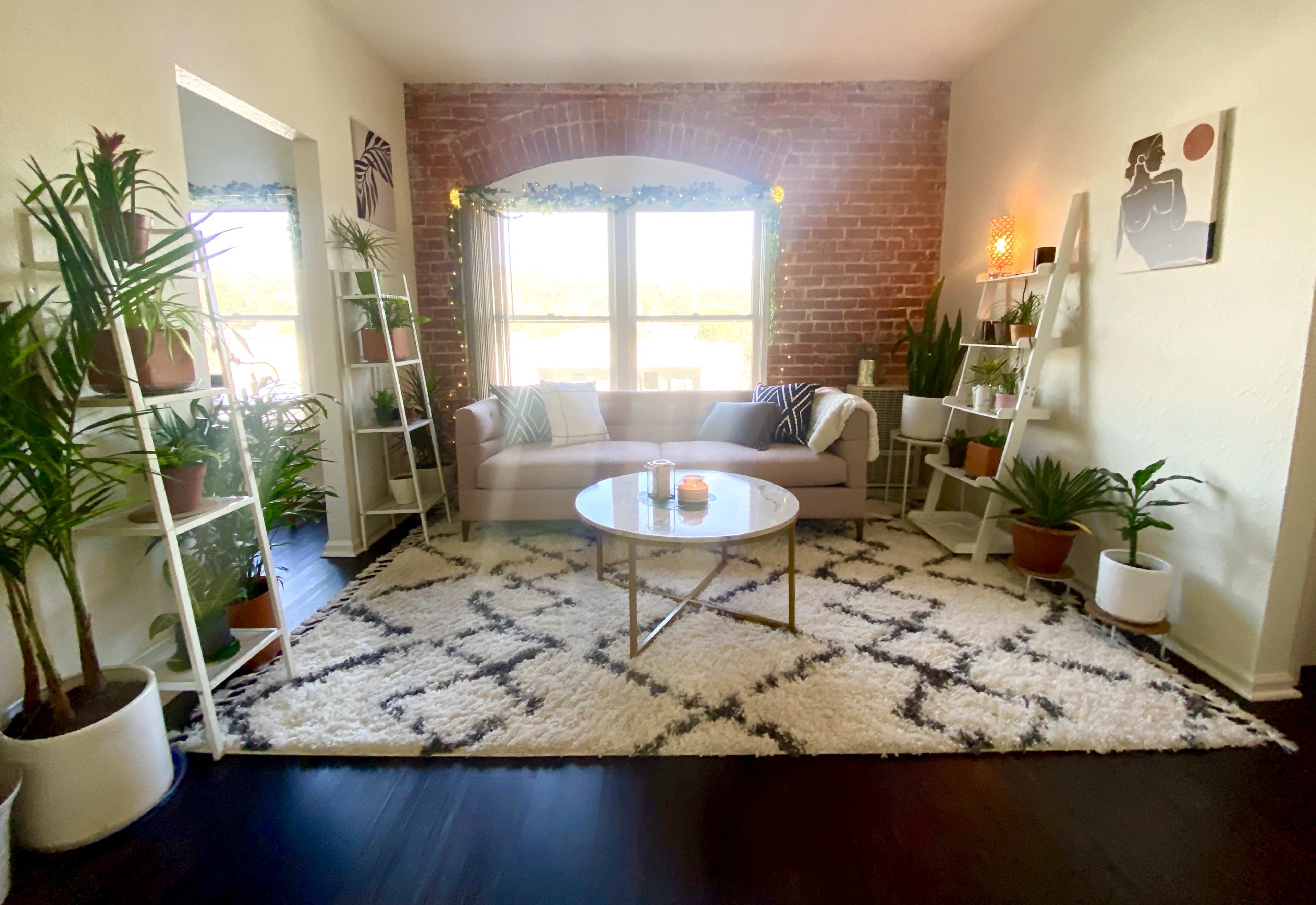 A cozy living room featuring a pink sofa, a round coffee table, numerous potted plants, and exposed brick walls.