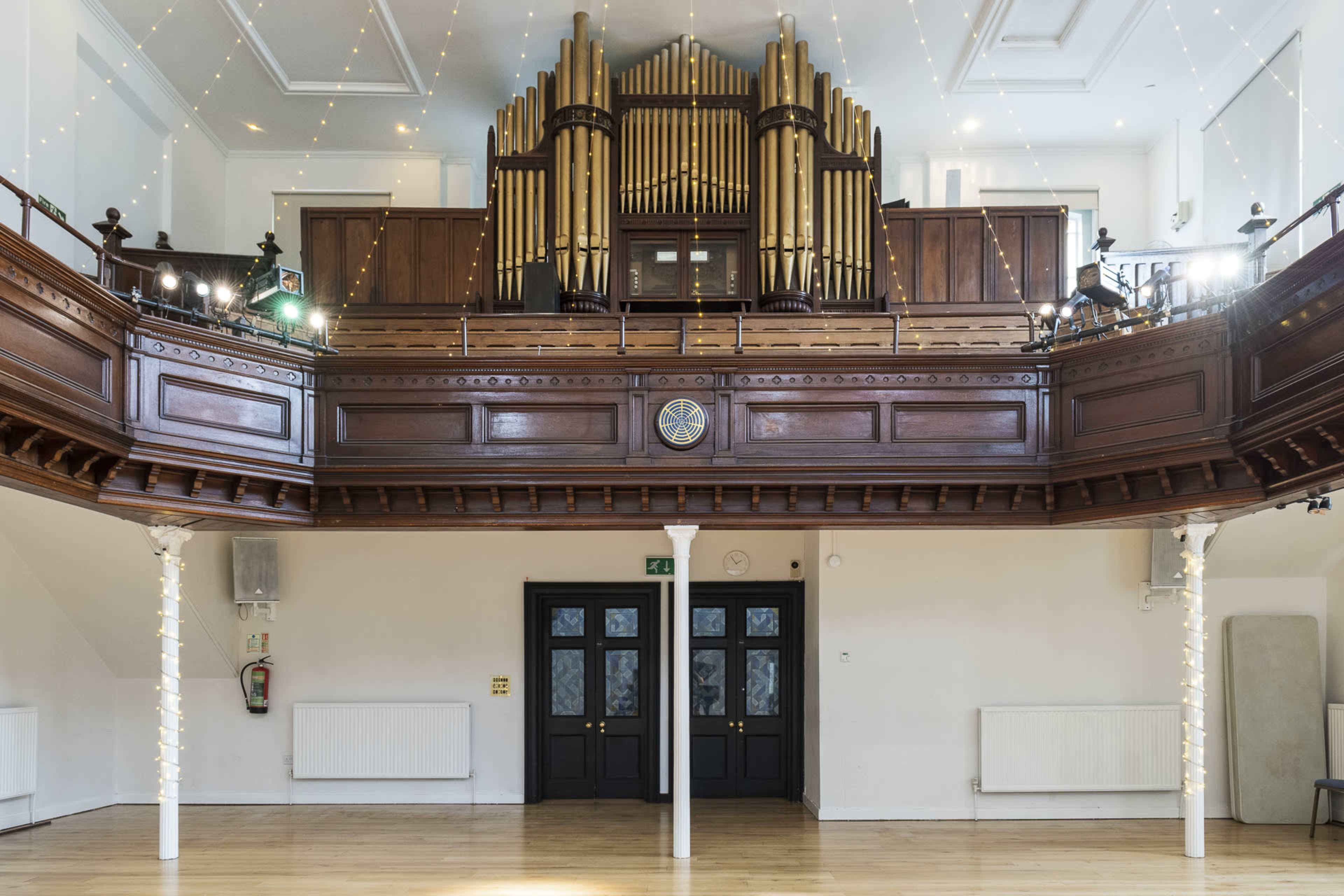 A spacious hall featuring a large organ on an elevated platform, with wooden paneling, and two sets of double doors at the front.