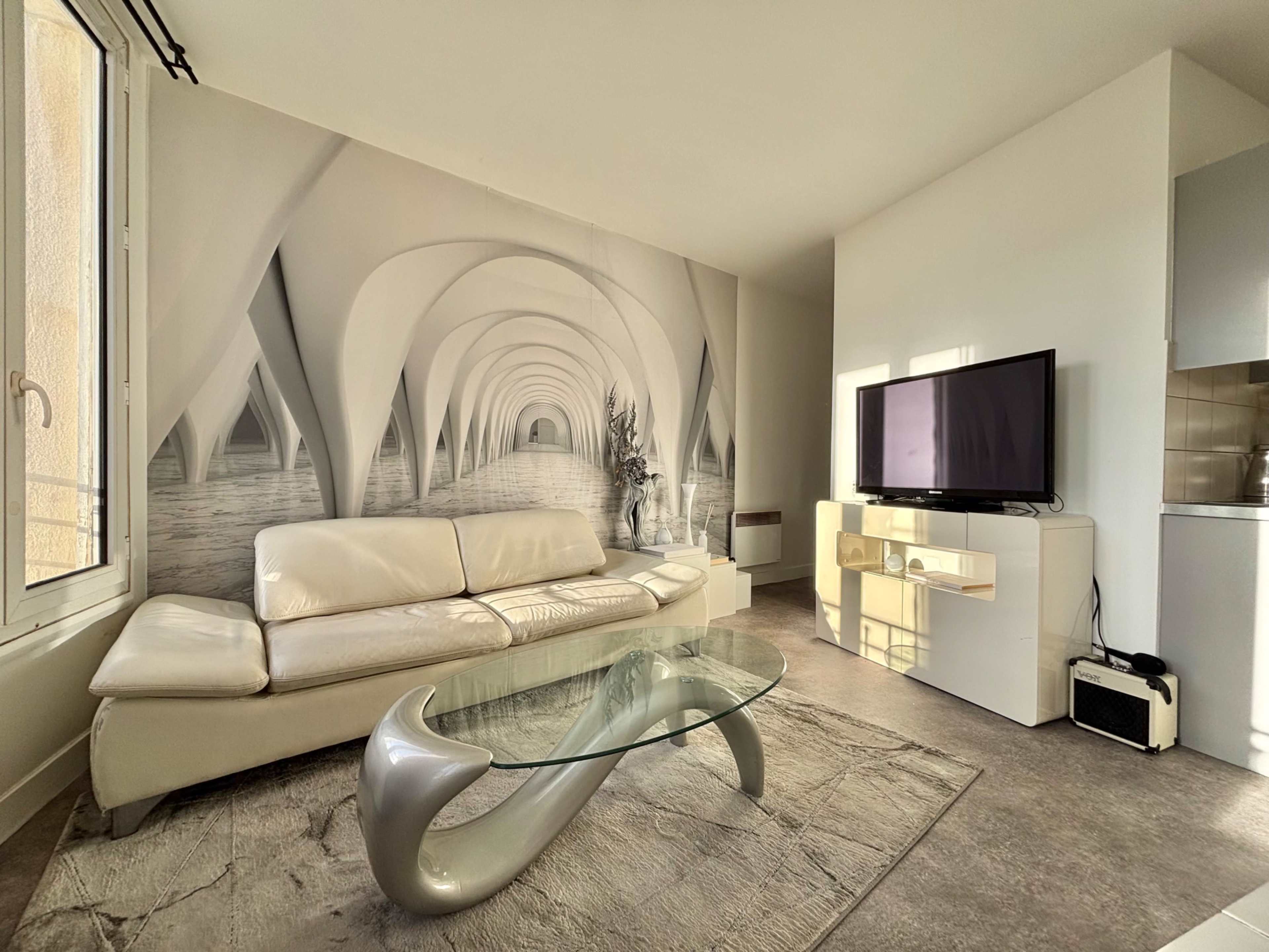 The image shows a modern living room with a light-colored sofa, a glass coffee table, a large television on a white console, and a mural featuring an abstract architectural design on one wall.