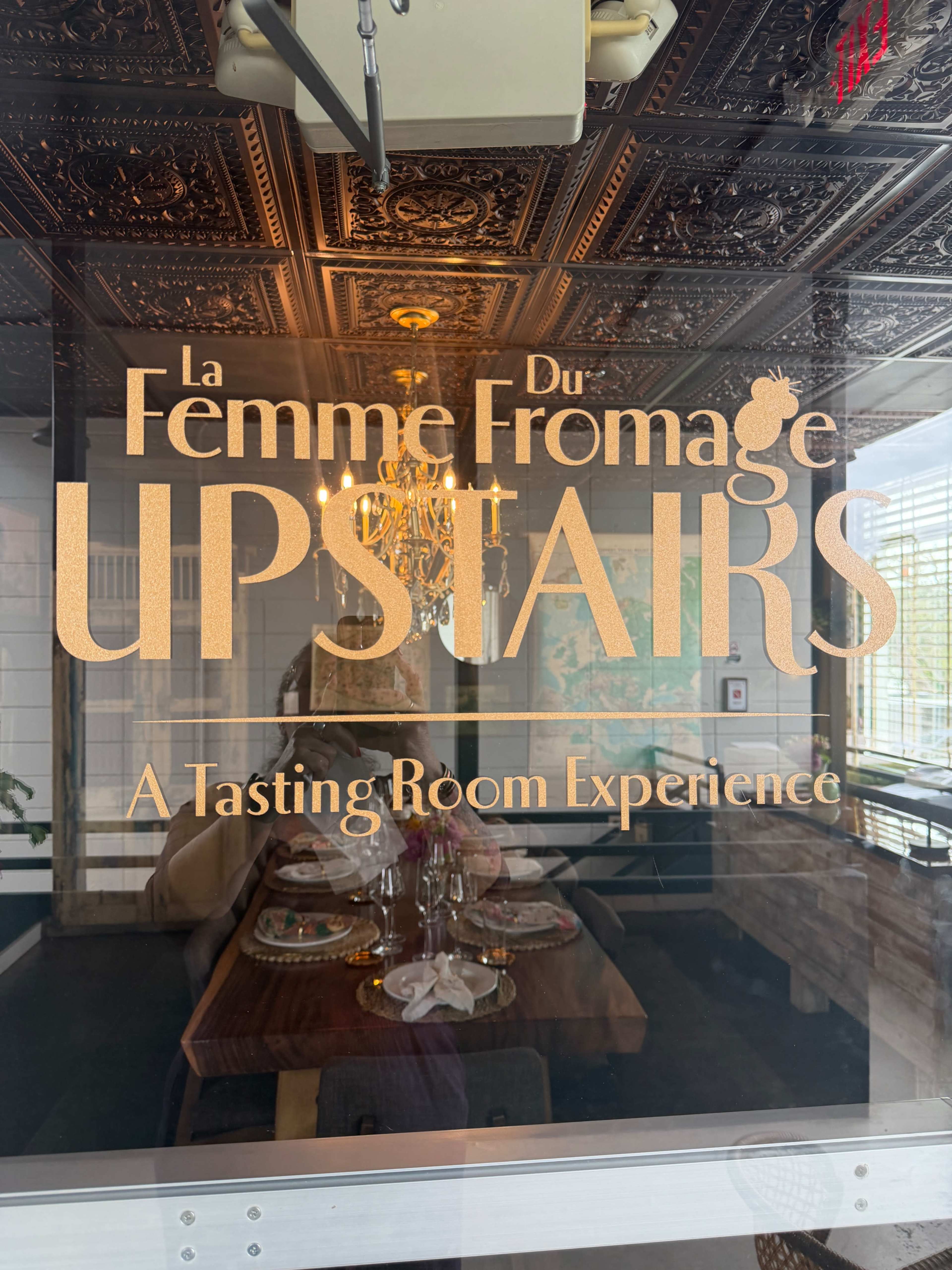 The image features a sign that reads "La Femme Du Fromage UPSTAIRS" and "A Tasting Room Experience," displaying an elegant font against a glass door.