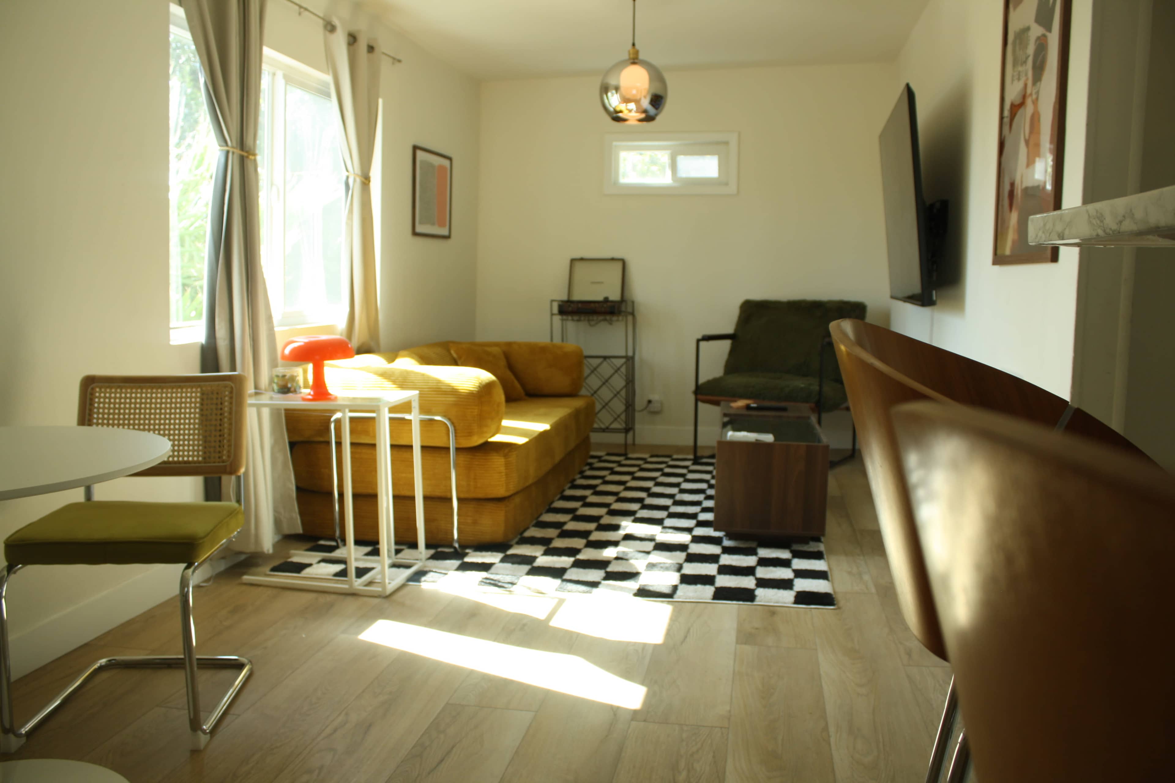 The image shows a modern living room with a yellow sofa, a green chair, a round table, and large windows allowing natural light to enter.