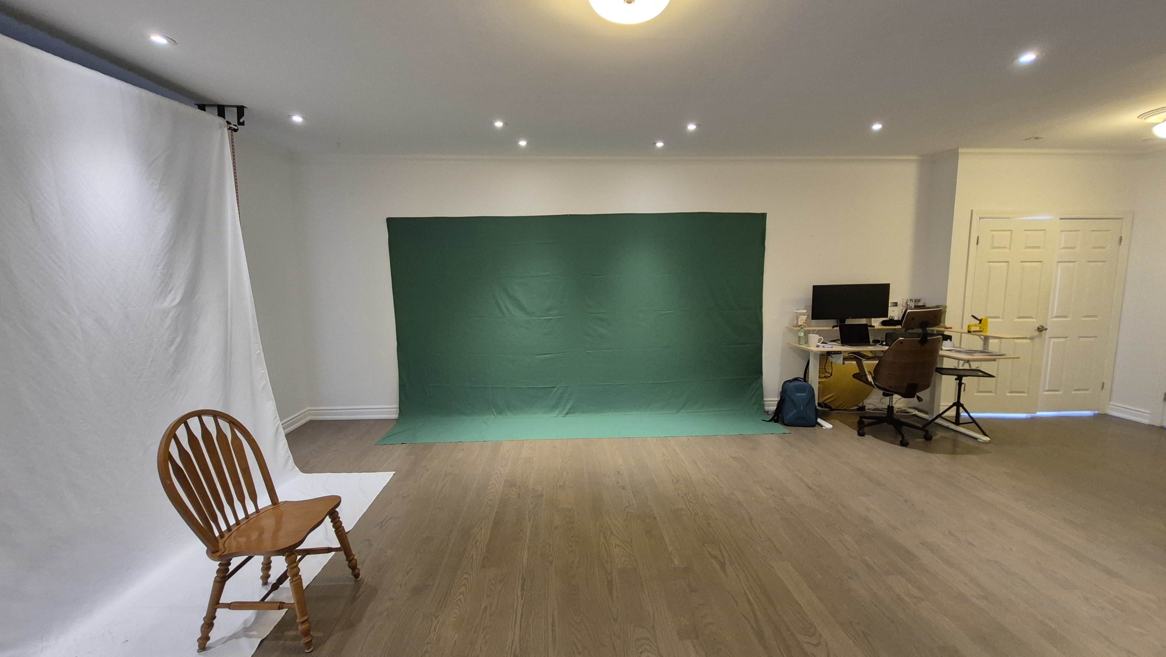 The image shows a spacious room featuring a green backdrop and a white backdrop, a wooden chair, and a desk with a computer and office chair.