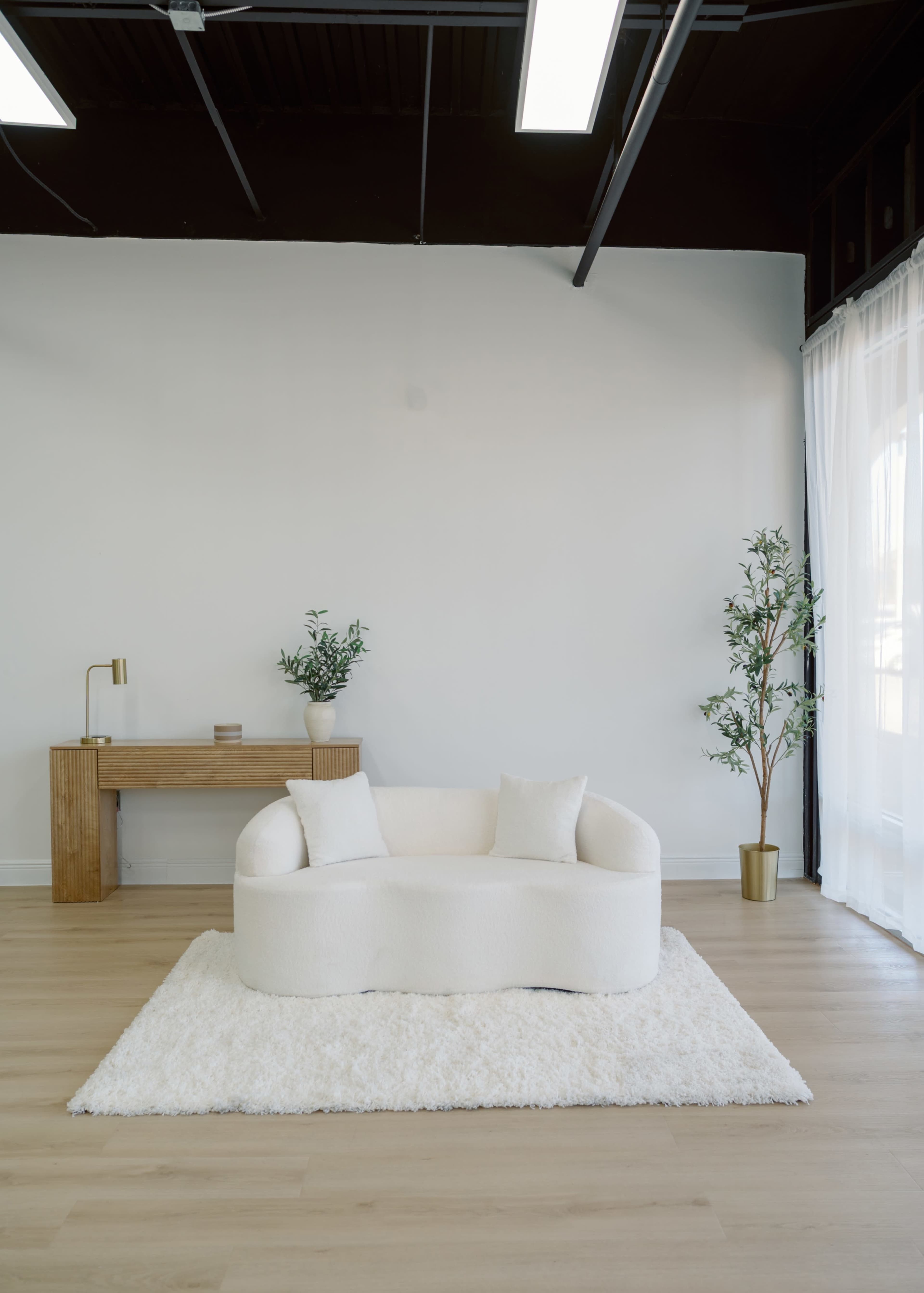 The scene features a white, curved couch on a plush rug, accompanied by a wooden desk and indoor plants, set against a light-colored wall.