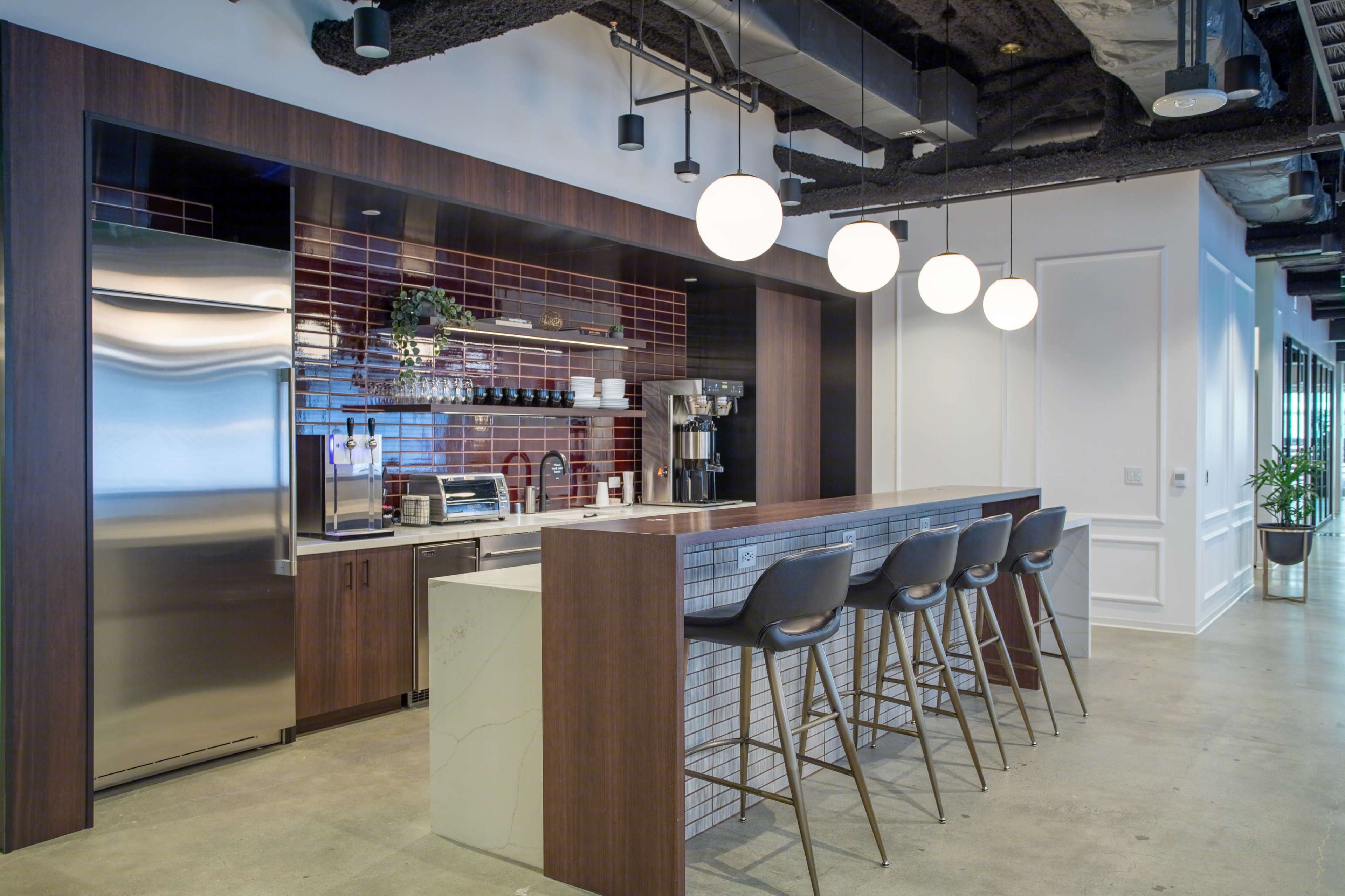 A modern office kitchen area featuring a stainless steel refrigerator, a coffee machine, and a bar with four high stools under pendant lighting.