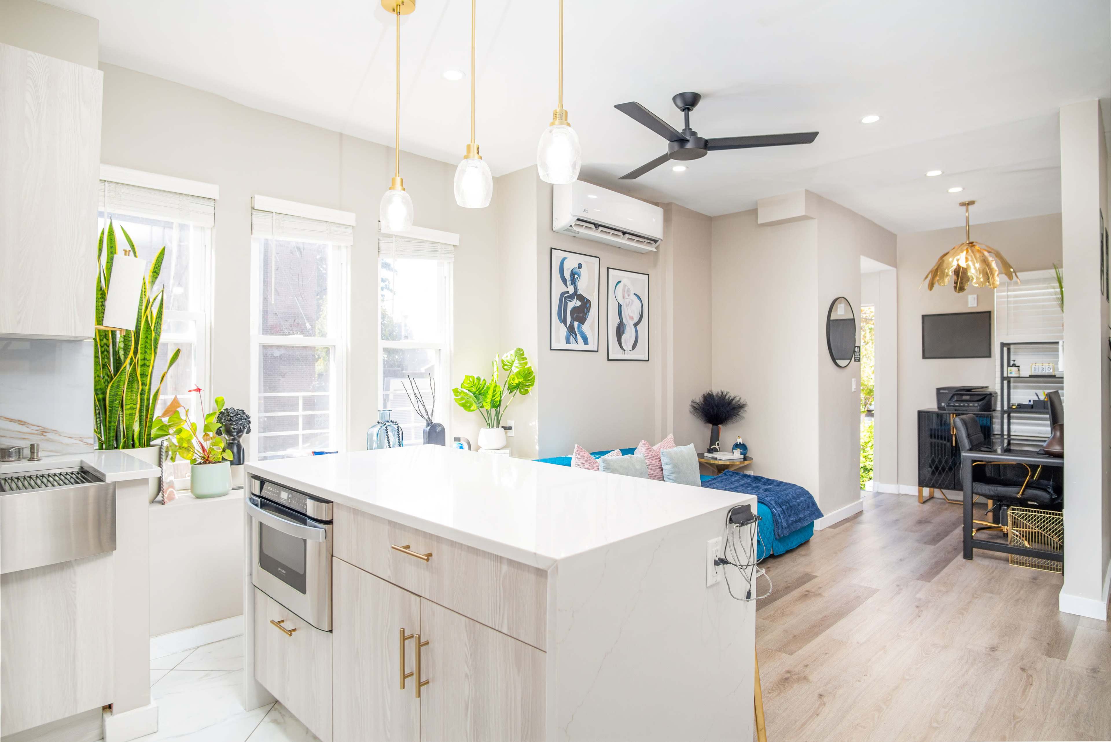 A modern kitchen with hanging light fixtures, an island, and an open layout leading to a living area with a blue couch and a desk space.