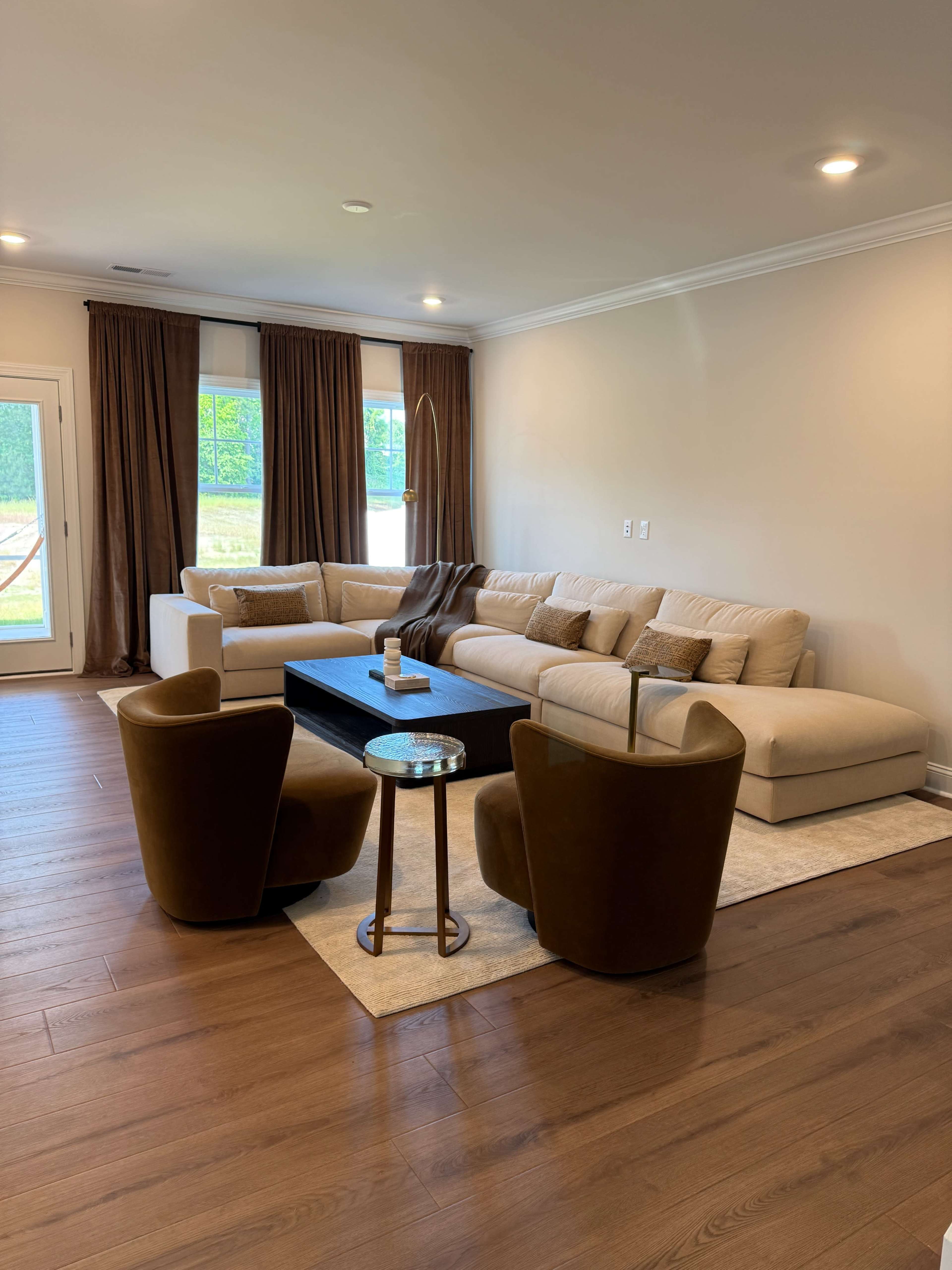 A light-colored sectional sofa is arranged around a black coffee table, complemented by two brown armchairs in a spacious living room with large windows and brown curtains.