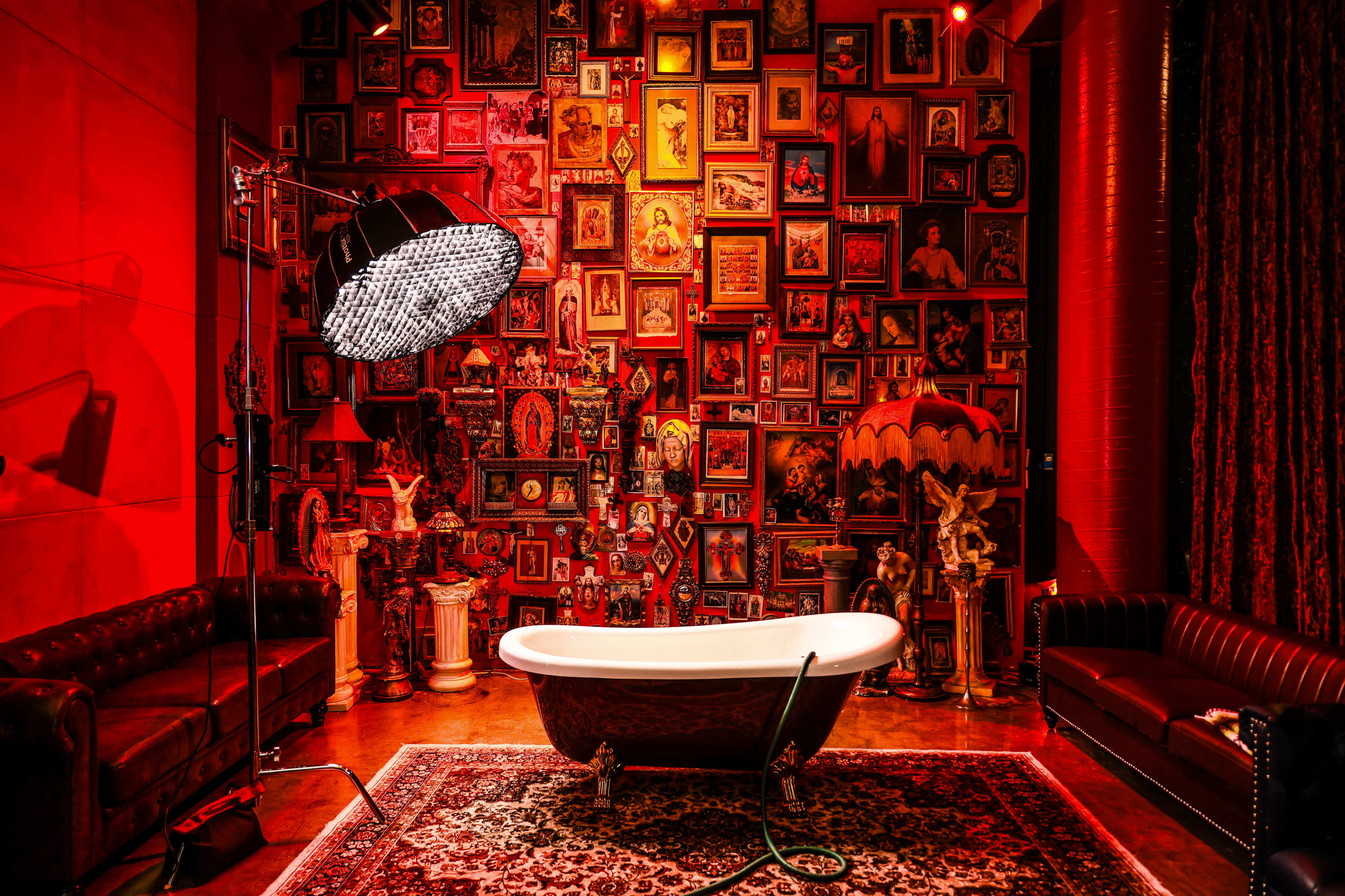 A luxurious bathroom featuring a vintage clawfoot tub, surrounded by an ornate wall filled with framed portraits and decorative objects, illuminated by a large studio light.