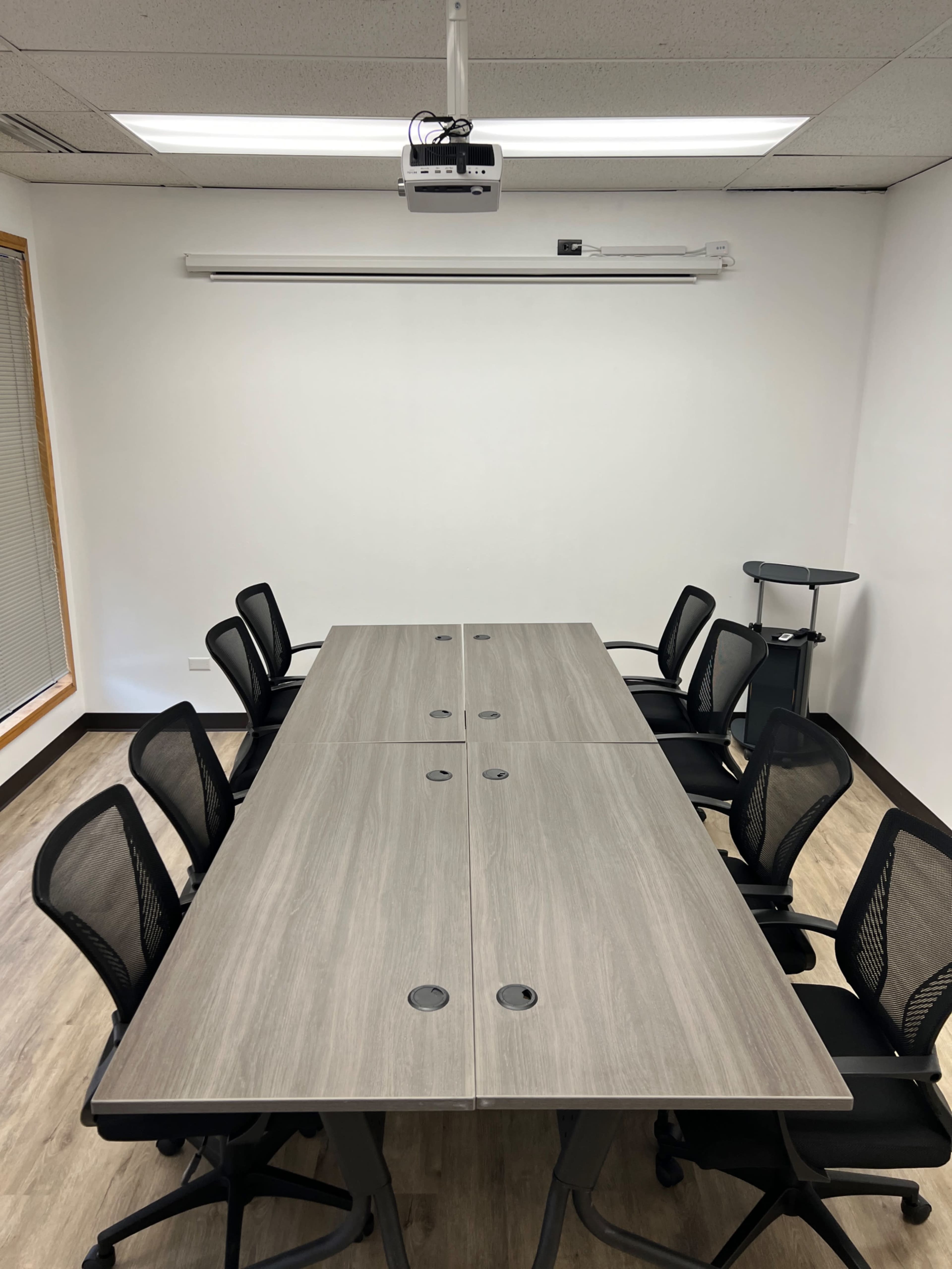 A rectangular conference table with eight black chairs is positioned in a brightly lit room featuring a wall-mounted projector.