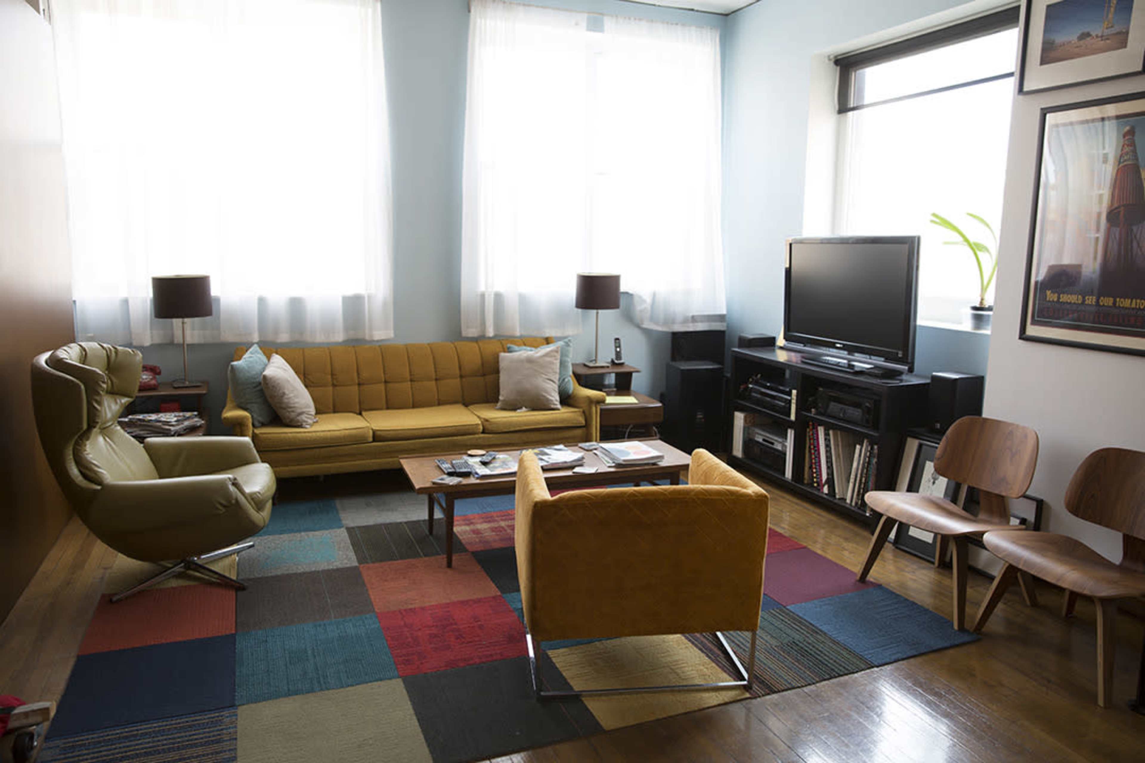 A living room with a yellow sofa, an olive green armchair, a wooden coffee table, a television, and hardwood floors, illuminated by natural light through large windows.