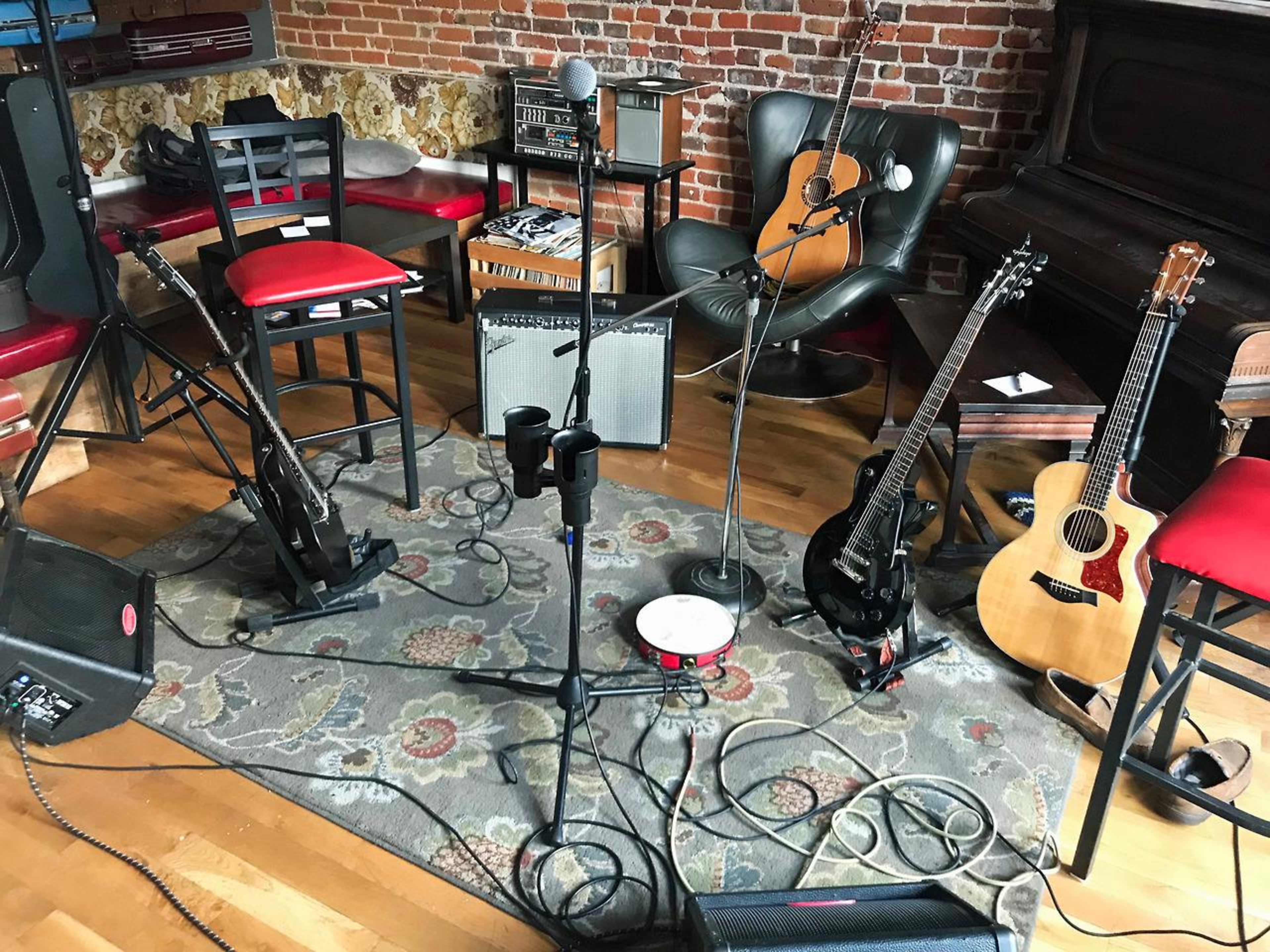A music setup with microphones, guitars, an amplifier, and stools arranged on a patterned rug in a cozy room with exposed brick walls.
