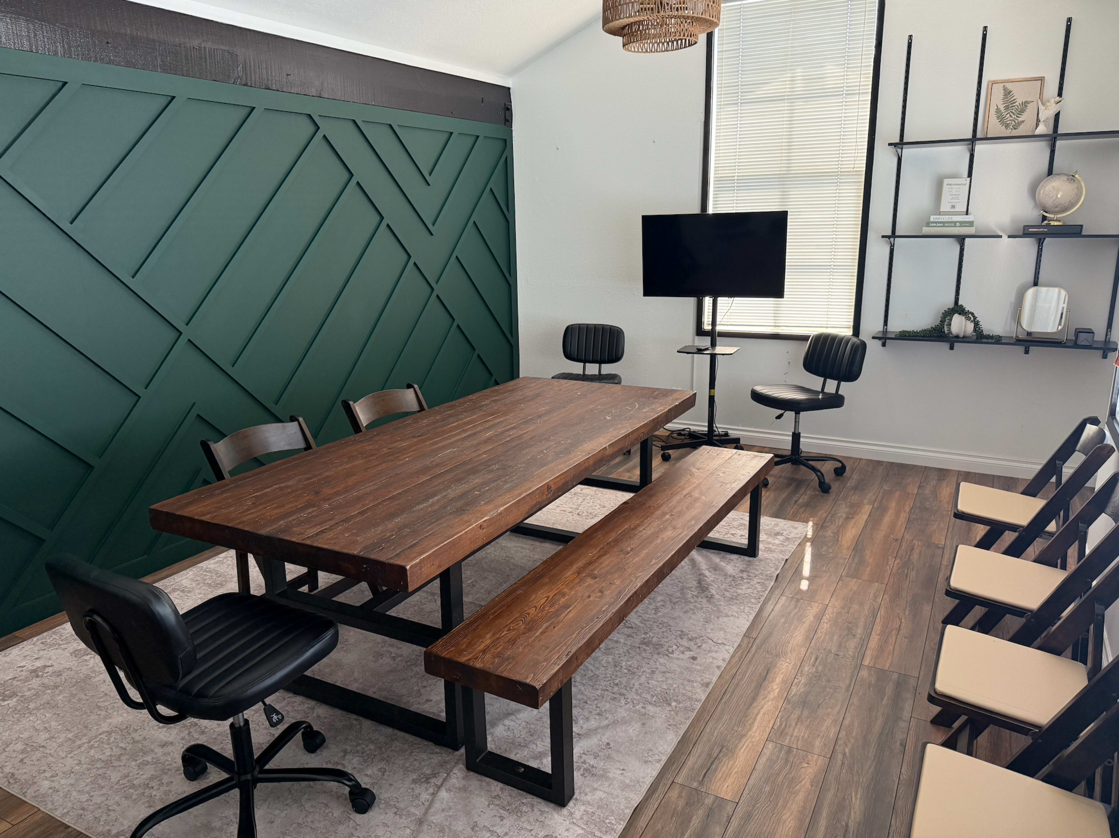 The image shows a meeting room featuring a large wooden table with benches and chairs, a television on a stand, and a green accent wall.