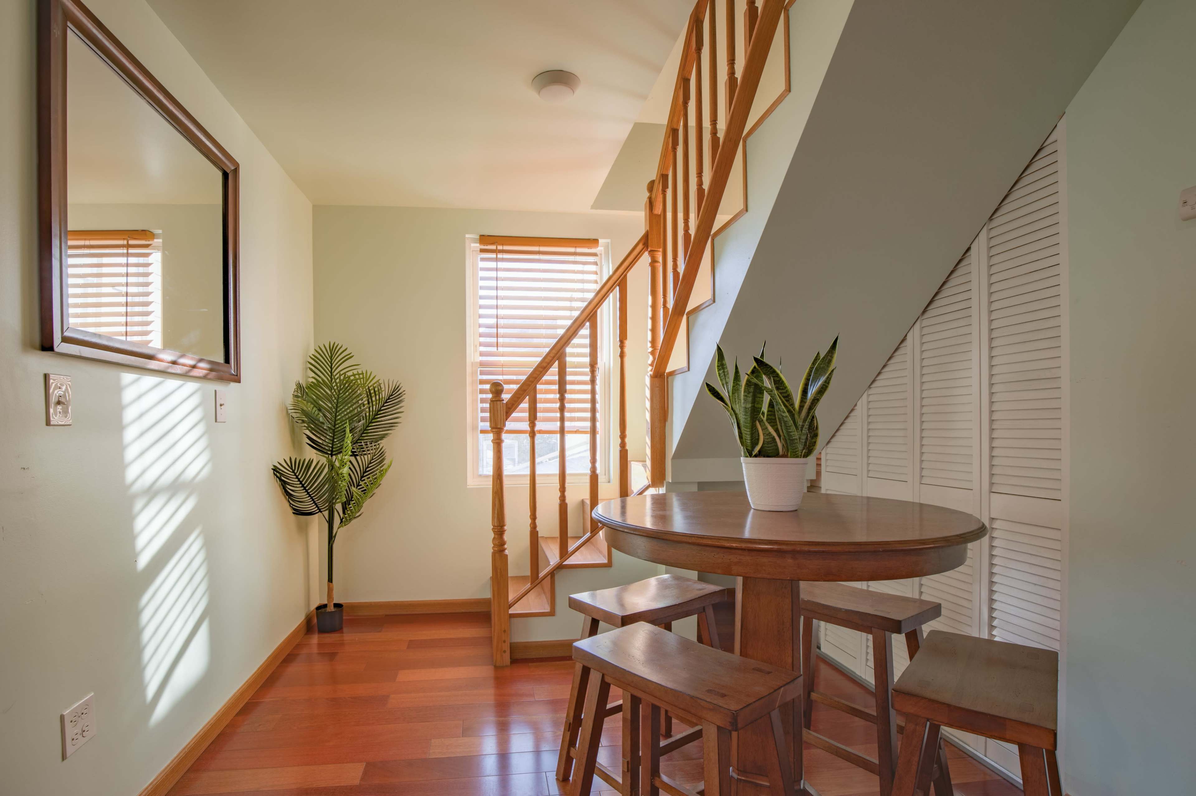 The image shows a small dining area with a round wooden table and four wooden chairs, adjacent to a staircase and featuring a plant near a window with wooden blinds.