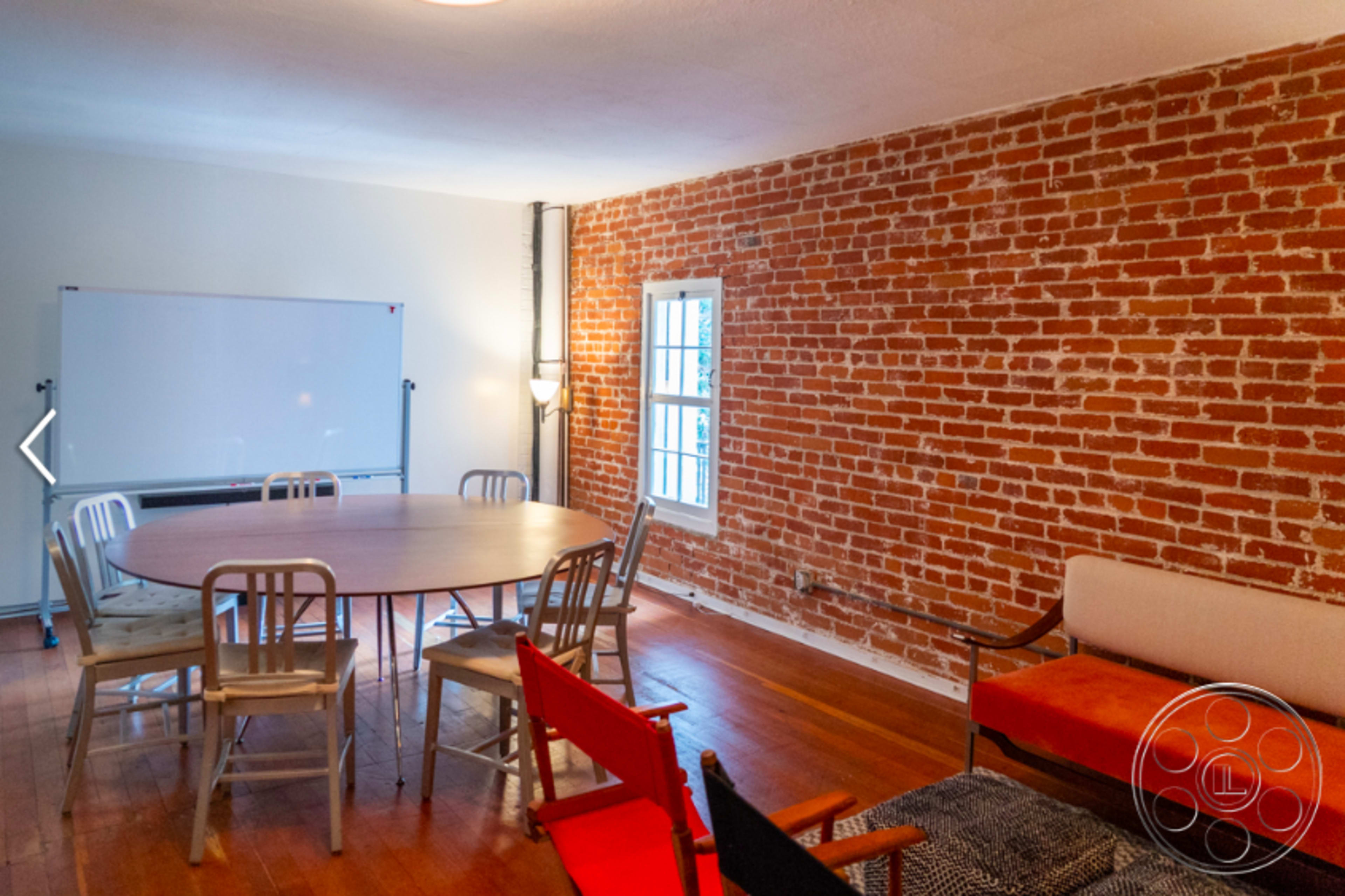 The image shows a meeting room with a large wooden table surrounded by chairs, brick walls, and a whiteboard on one side.