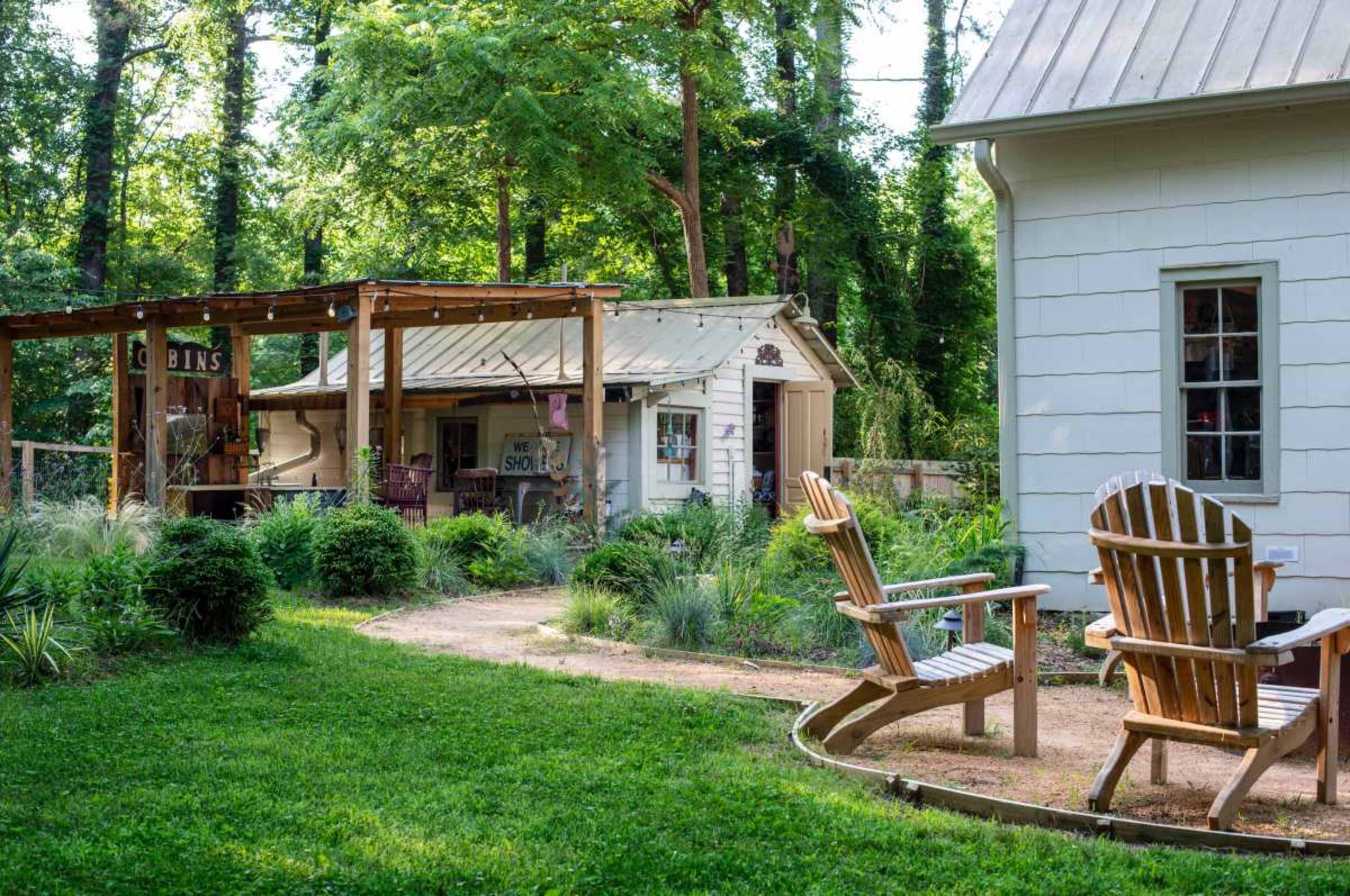 A winding path leads through a lush garden to a small cabin and an adjacent house, with two wooden chairs placed on the grass.