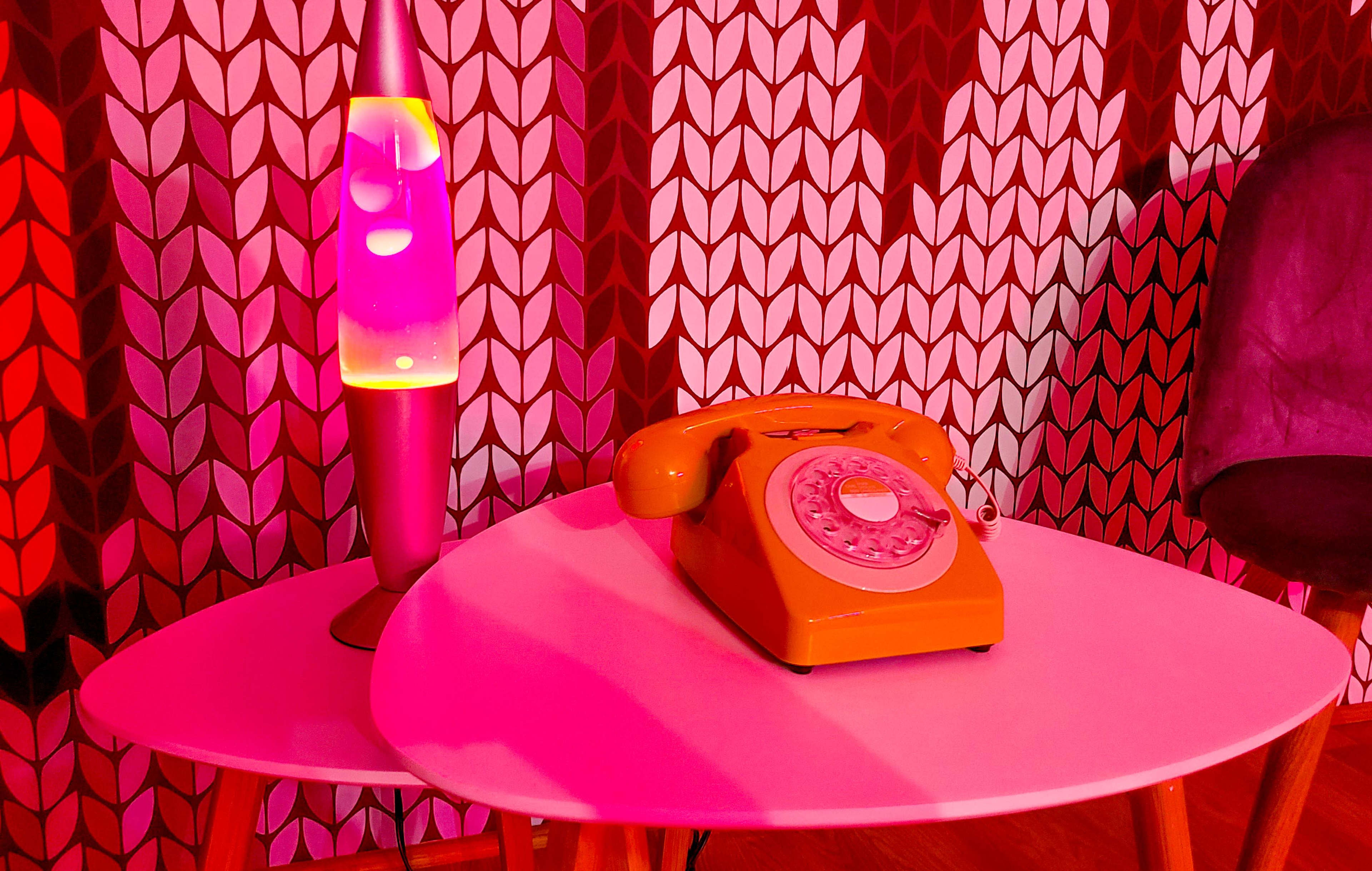 An orange rotary telephone rests on a white table next to a purple lava lamp, set against a patterned background.