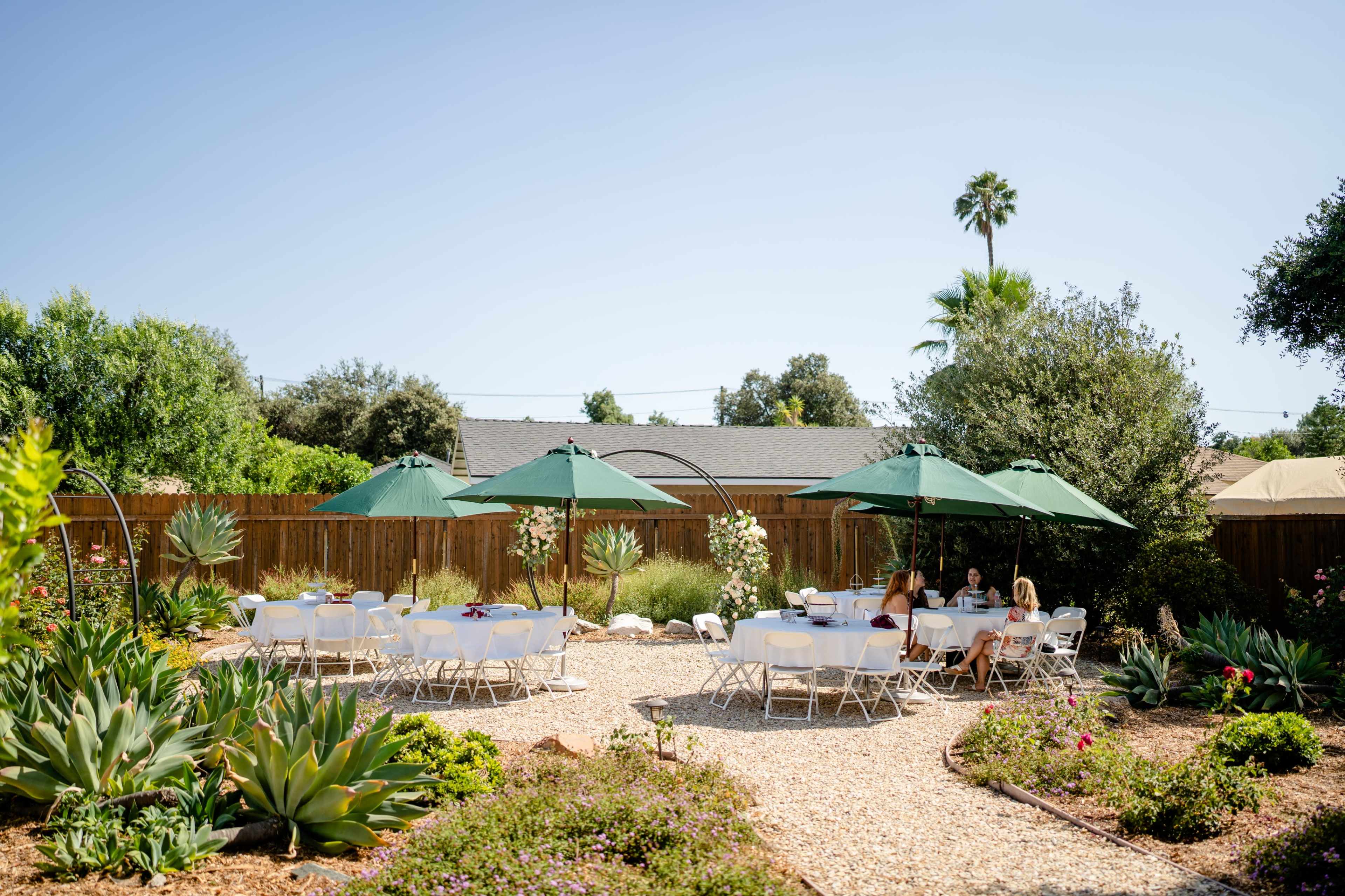 A garden area features several white tables and green umbrellas arranged on gravel amid lush plants and trees.