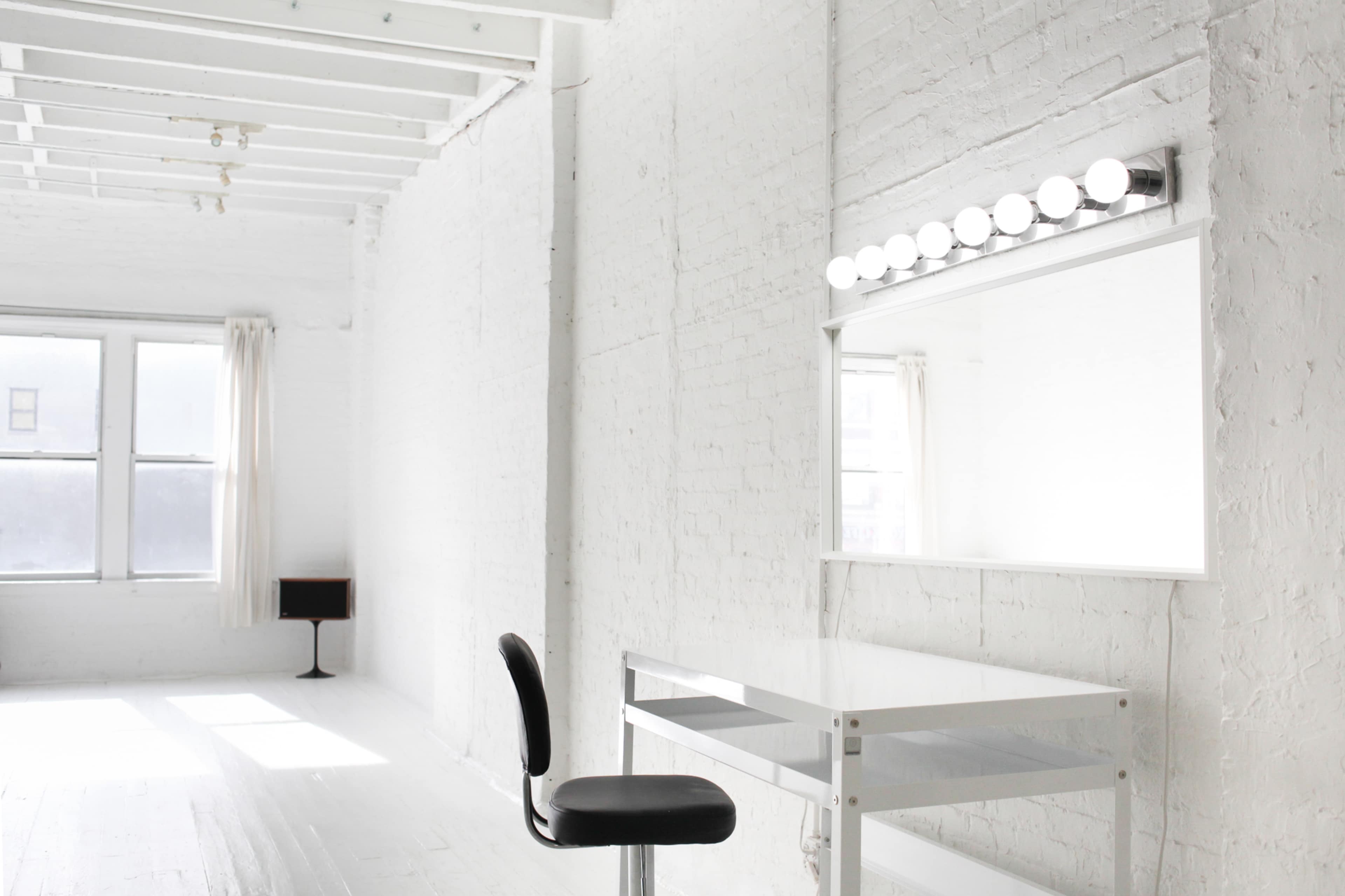 A bright, minimalist room with white walls, a large mirror with lights, a desk, and a chair.