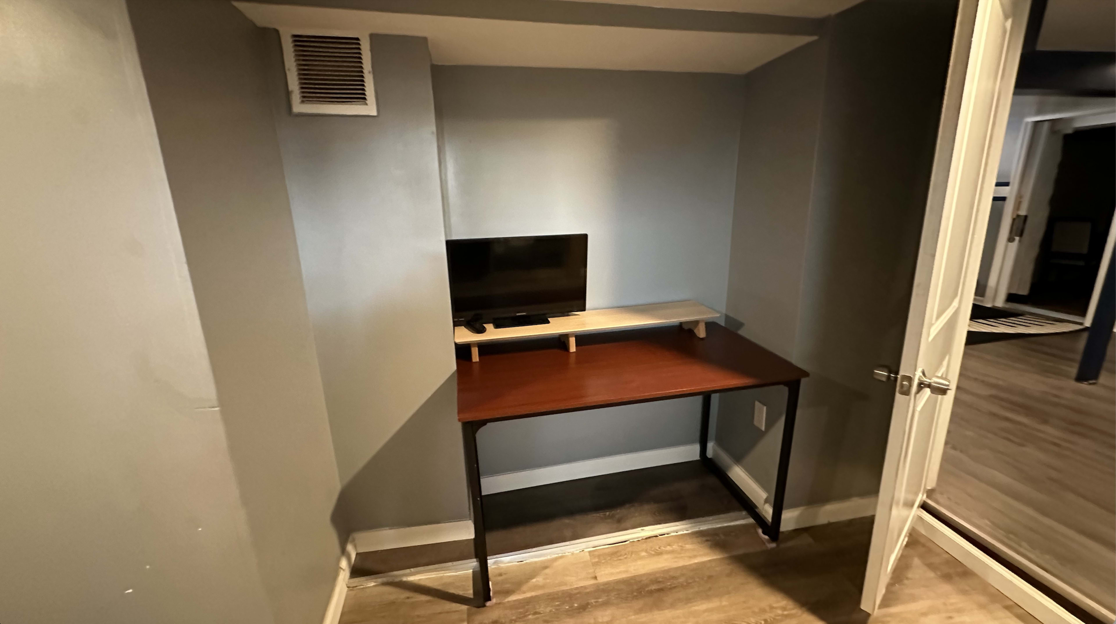 A small workspace featuring a wooden desk with a monitor and a shelf, positioned in a corner of a room with gray walls.