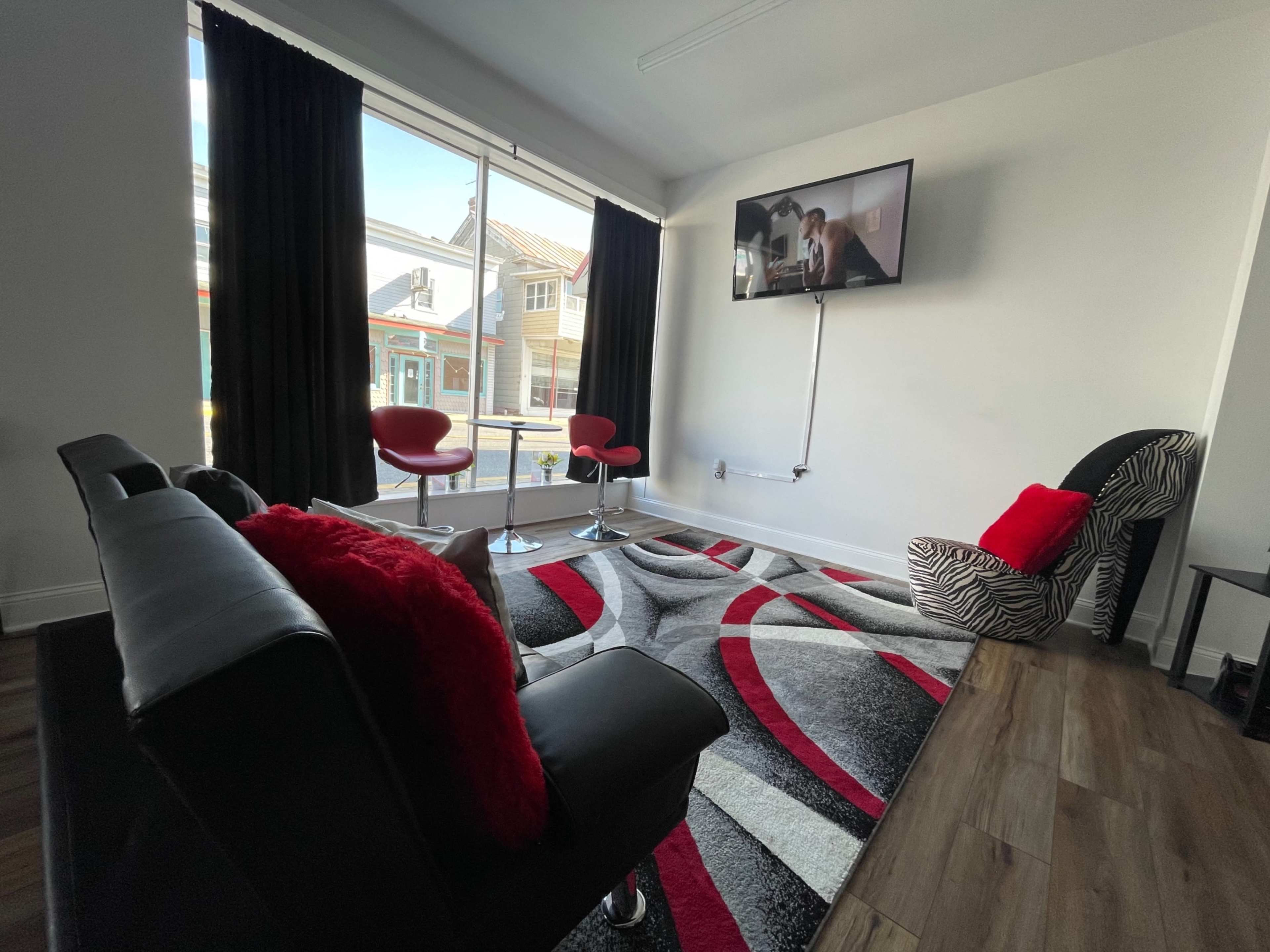 A modern living room with a large window, a black sofa, red and white decor, and two bar stools facing a wall-mounted television.