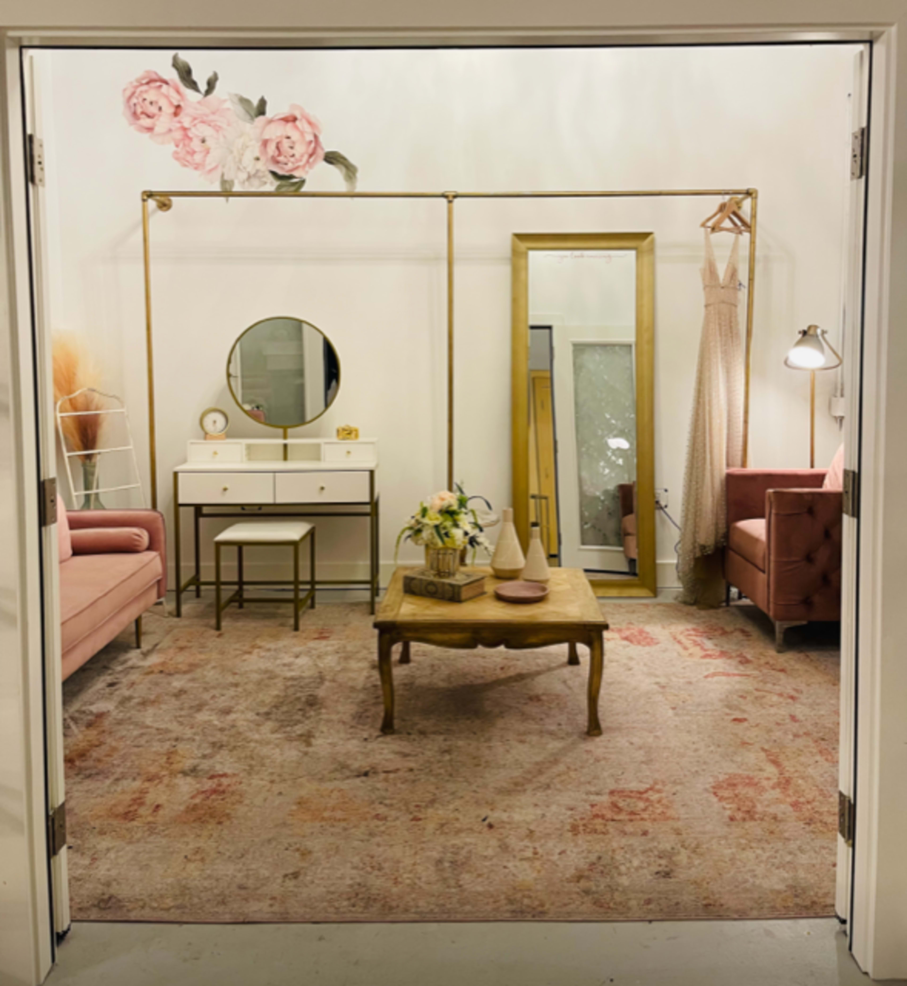 A styled room with a pink sofa, a beige armchair, a wooden coffee table, a round mirror, and a large mirror against the wall, surrounded by floral decor.