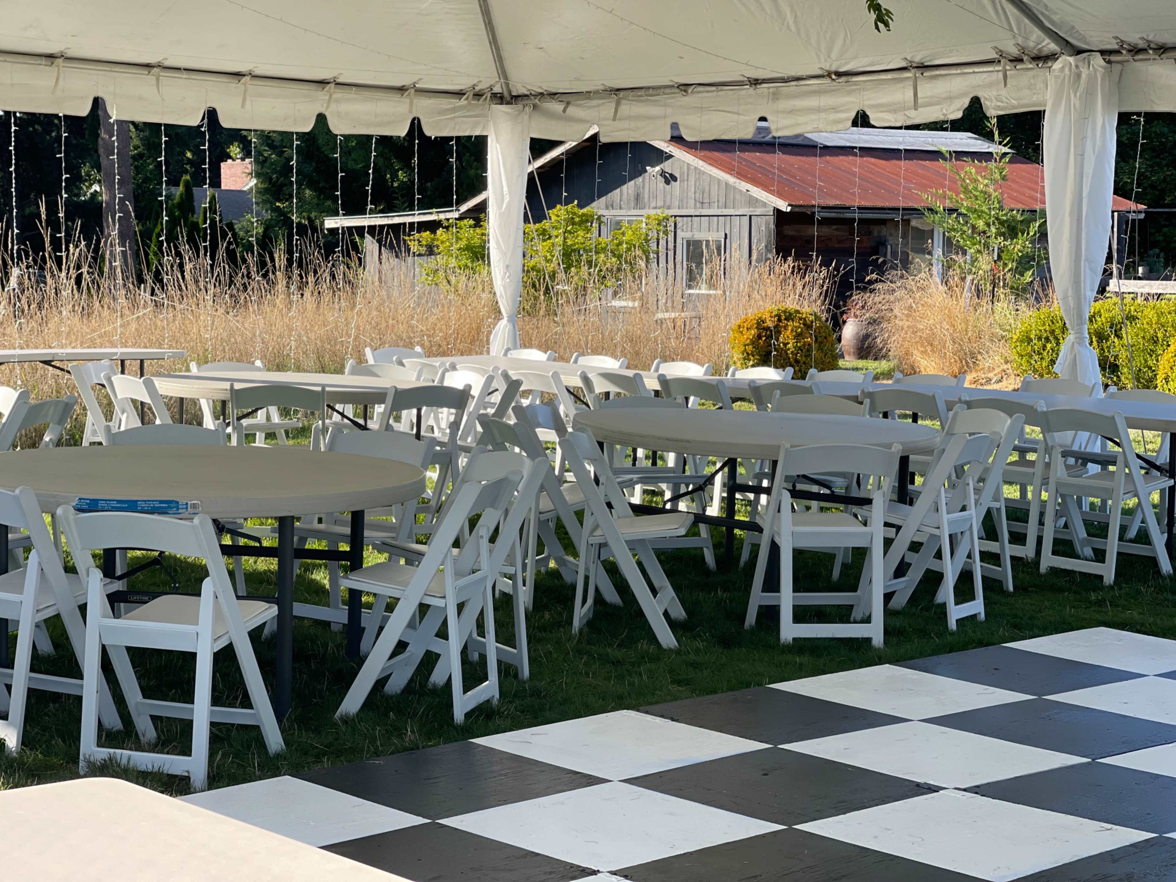 A tent is set up with several round tables and white chairs arranged on a black and white checkered floor, overlooking a grassy area with a rustic building in the background.