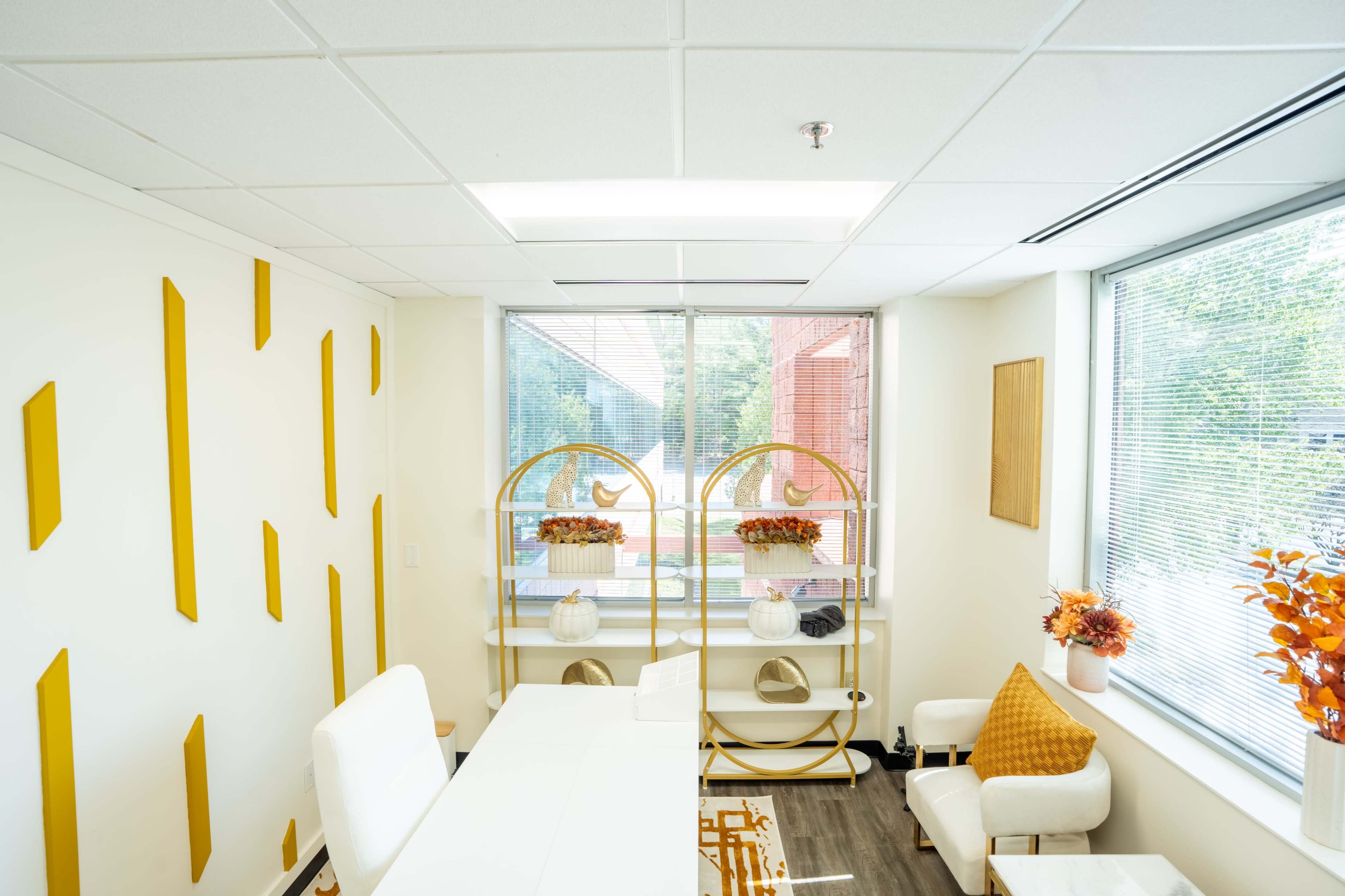 The image shows a modern office space featuring white walls, a desk, a shelf with decorative items, and large windows allowing natural light to enter.